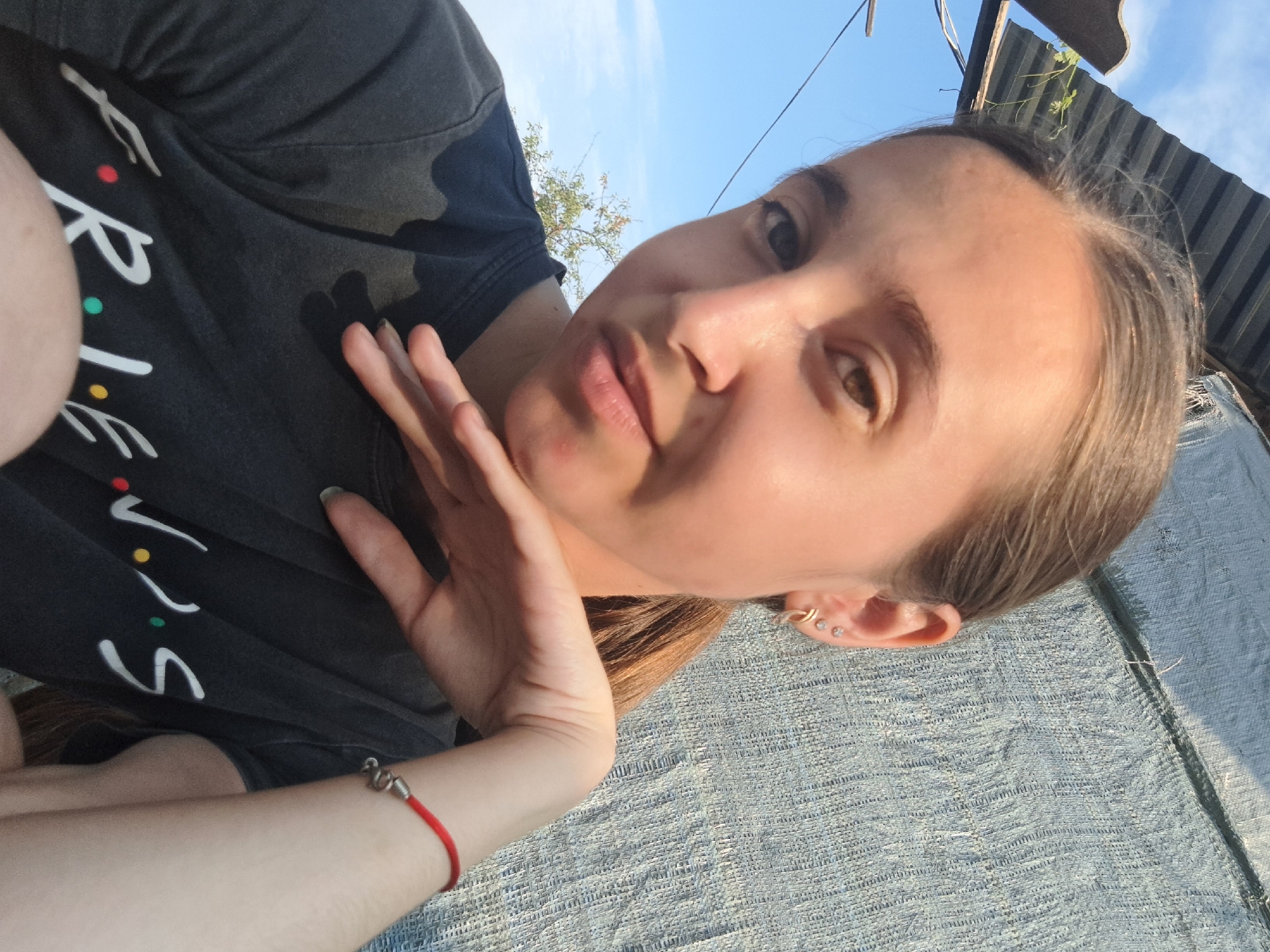A young woman with light brown hair pulled back in a ponytail, wearing a dark gray t-shirt with the "FRIENDS" logo visible, is captured in a close-up, head-and-shoulders selfie. She has light skin, brown eyes, and is wearing multiple earrings in her left ear. Her left hand is resting on her chin, and a red string bracelet with a silver snake charm is visible on her right wrist. The background is a textured blue-gray tarp or fabric, with a corrugated metal roof and blue sky visible in the upper left corner, suggesting an outdoor setting, likely in Vypasne, Ukraine, given the context. Sunlight casts a shadow on the left side of her face and shoulder.