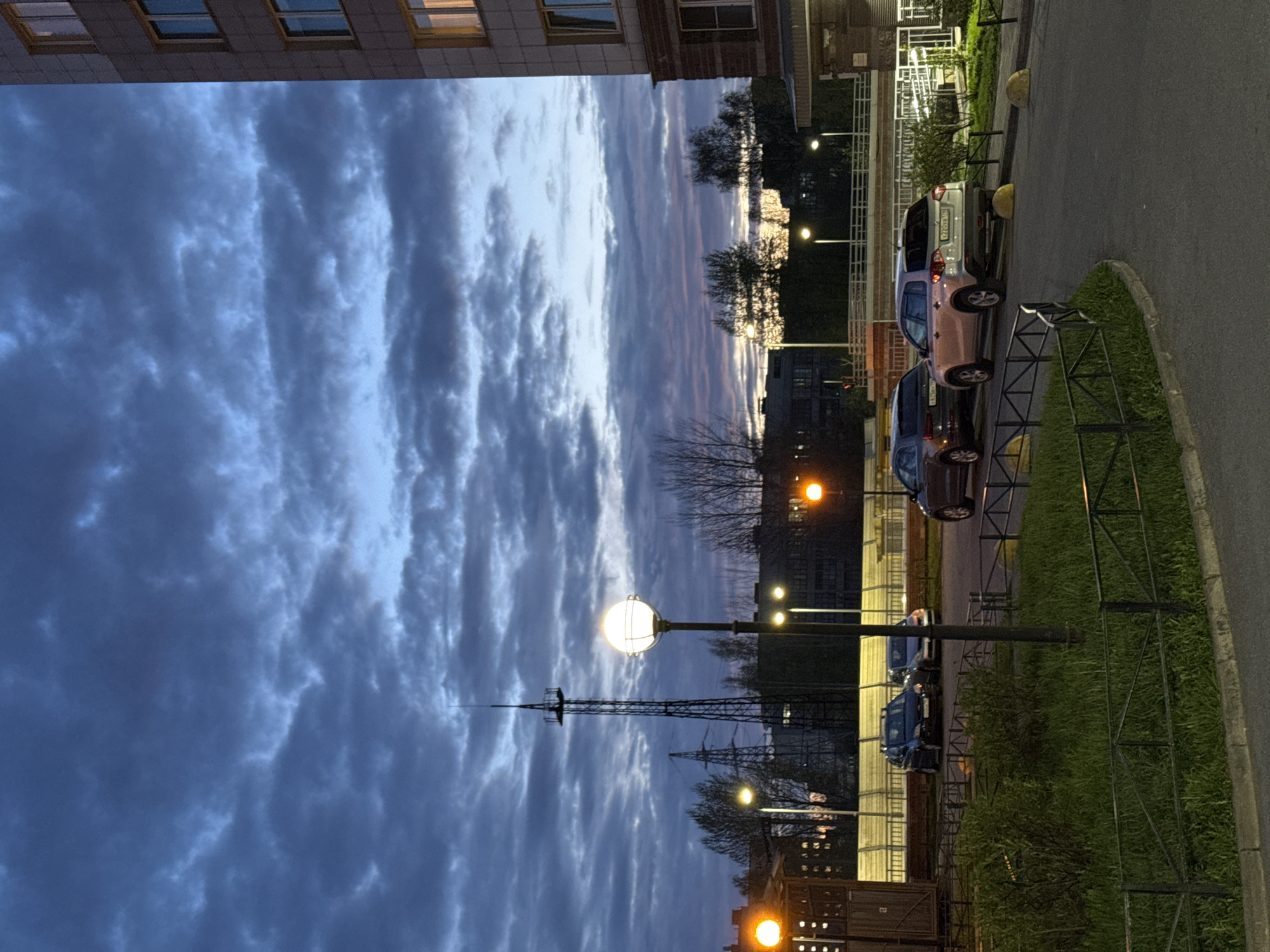 The image depicts a street scene in Saint Petersburg, Russia, at dusk or dawn. The sky is filled with striated clouds, tinged with hues of blue, gray, and hints of pink. The architecture includes a modern apartment building on the upper left. A prominent telecommunications tower is visible against the skyline. Streetlights illuminate the scene. Several parked cars line the right side of the road along the sidewalk. A low metal fence separates the sidewalk from a narrow strip of grass. The photograph captures a quiet moment in the urban landscape.