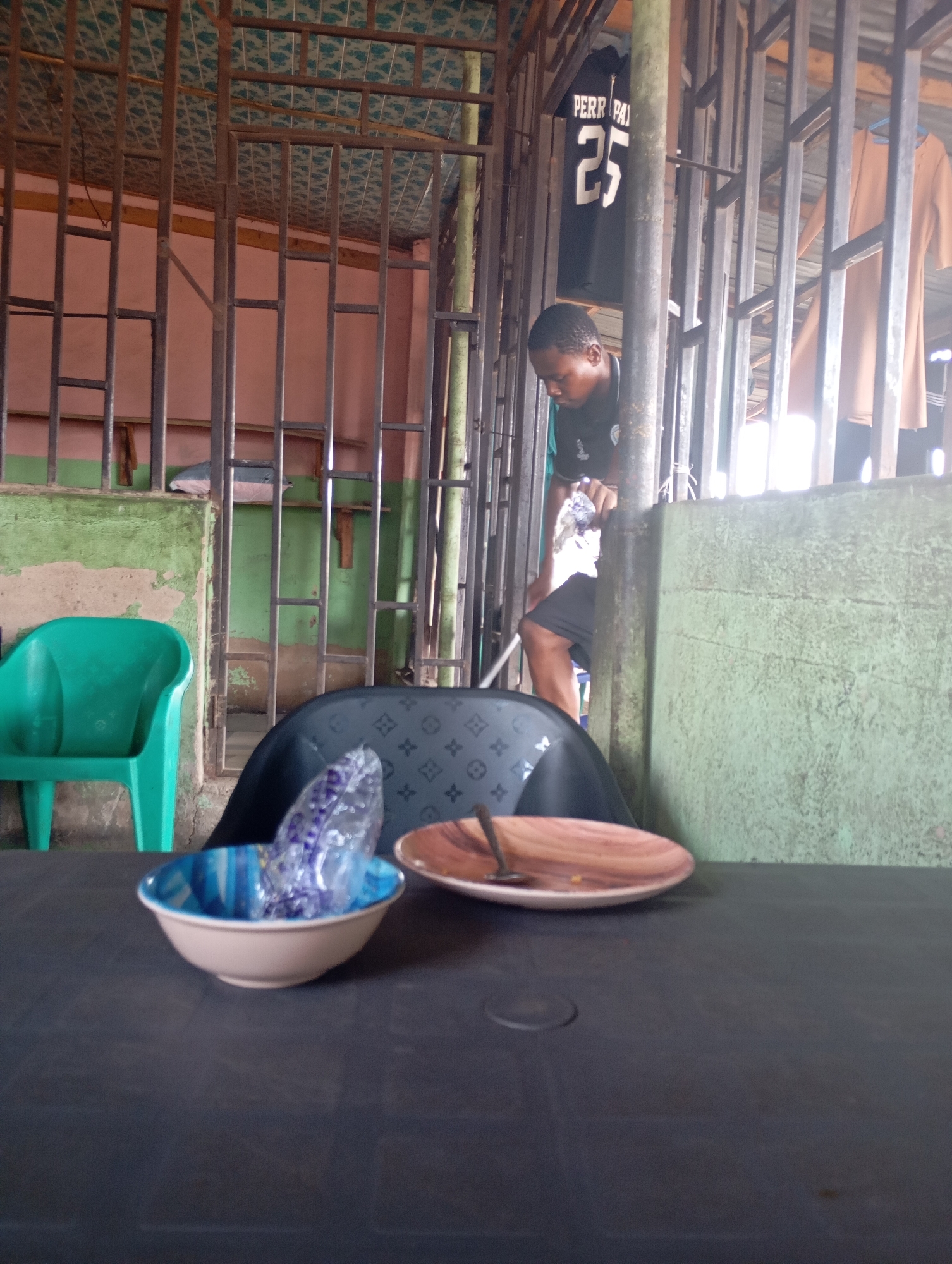 In a dimly lit, open-air structure with a barred metal facade, a young man is captured in the act of placing a clear plastic bag into a bowl. The man, wearing a dark t-shirt with a white logo and dark shorts, stands to the right of the frame, his attention focused on his task. Behind him, a black t-shirt with the number "25" and the word "PERR-PAI" is visible, hanging amongst other clothing items.
The foreground of the image is dominated by a dark, rectangular table. On the table, a light-colored bowl with blue and white interior patterns sits to the left, containing the plastic bag. To its right, a brown, shallow dish with a small spoon rests. A green plastic chair is positioned on the left side of the frame, while a dark chair with a patterned back is behind the table. The background consists of green and pink painted walls, interspersed with horizontal wooden beams and a patterned ceiling. The overall impression is of a casual, possibly communal dining or gathering space, typical of informal eateries or hangouts in Shendam, Nigeria.