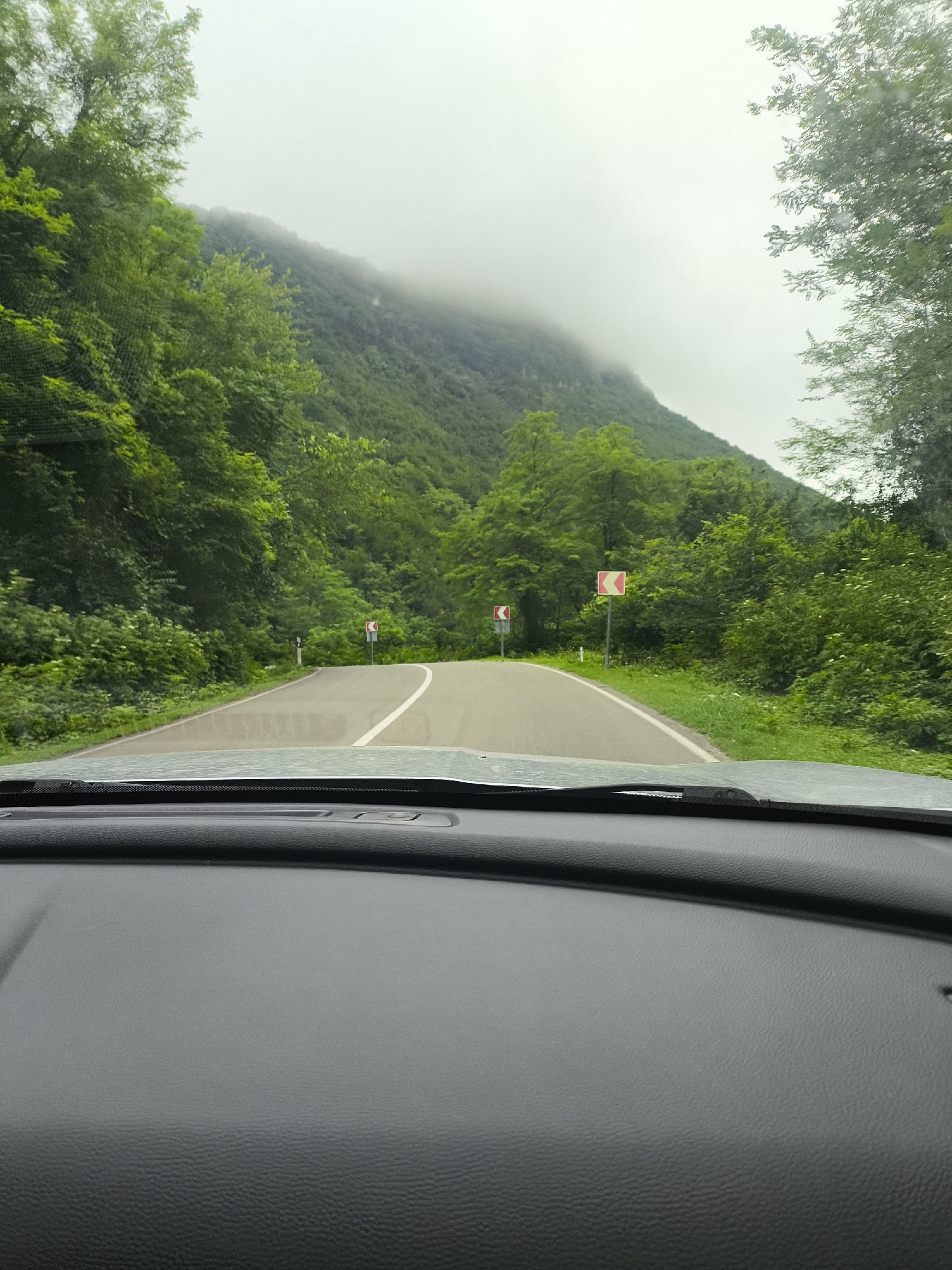 The image depicts a road winding through a lush, green mountainous landscape, possibly in the city of Gamoghma Nogha, Georgia. The road is paved with a white line dividing the lanes and is bordered by dense vegetation and tall trees on either side. Several road signs, indicating curves, are visible along the route. The sky is overcast and foggy, with mist obscuring the tops of the mountains in the distance. The perspective is from inside a vehicle, looking out through the windshield, with the dashboard visible in the foreground. The scene suggests a drive through a scenic, natural environment under a cloudy sky.