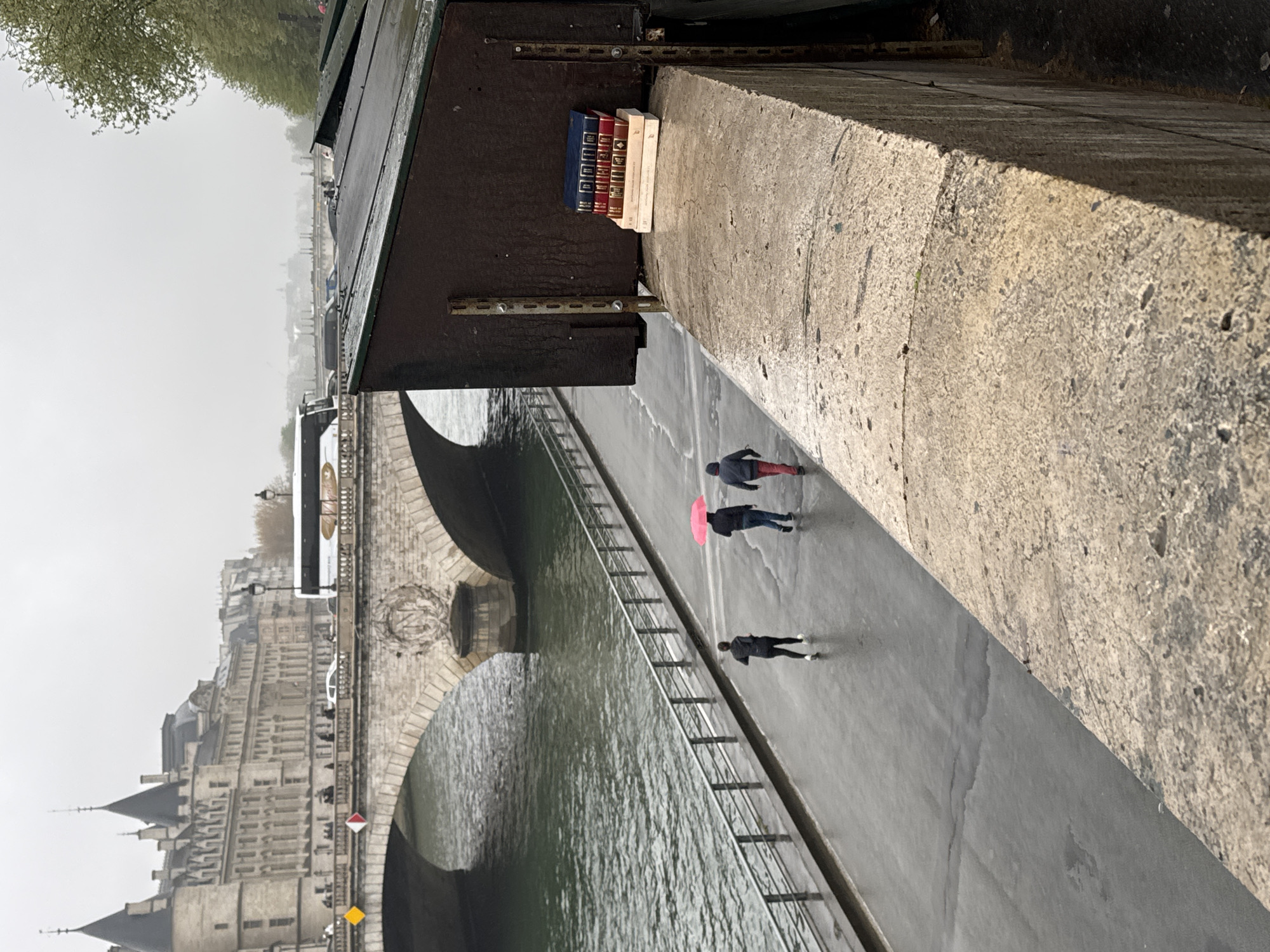 The image shows a section of the Seine River in Paris, France, with a view of buildings along the riverbank. A stone bridge with arches spans the river, and a pedestrian walkway runs alongside the water. Three individuals are walking on the pathway. One is holding a pink umbrella. A small book stall is attached to the bridge structure above the walkway. The day appears overcast and possibly wet, judging by the reflections on the walkway.