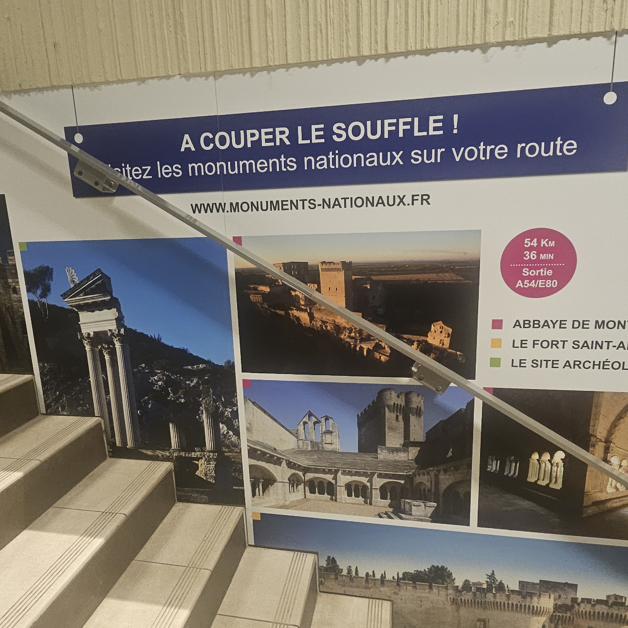This is a photograph of a promotional banner and posters displayed on a wall next to a staircase. The banner at the top reads "A COUPER LE SOUFFLE ! ...ez les monuments nationaux sur votre route" and "WWW.MONUMENTS-NATIONAUX.FR". Below the banner are several large photographic panels showcasing various historical sites. One panel features an ancient Roman ruin with columns against a blue sky. Another shows a medieval castle or fortress bathed in warm, low-angled light. A third displays a courtyard with arcades and a bell tower in the background. A pink circle on the right side of the banner indicates "54 Km 36 MIN Sortie A54/E80". Below this are color-coded labels: a purple square for "ABBAYE DE MON", a yellow square for "LE FORT SAINT-AN", and a green square for "LE SITE ARCHÉOL". The staircase in the foreground is made of beige tiled steps with textured treads. The overall impression is an advertisement for visiting national monuments, possibly located near Lançon-Provence, France, given the exit information.