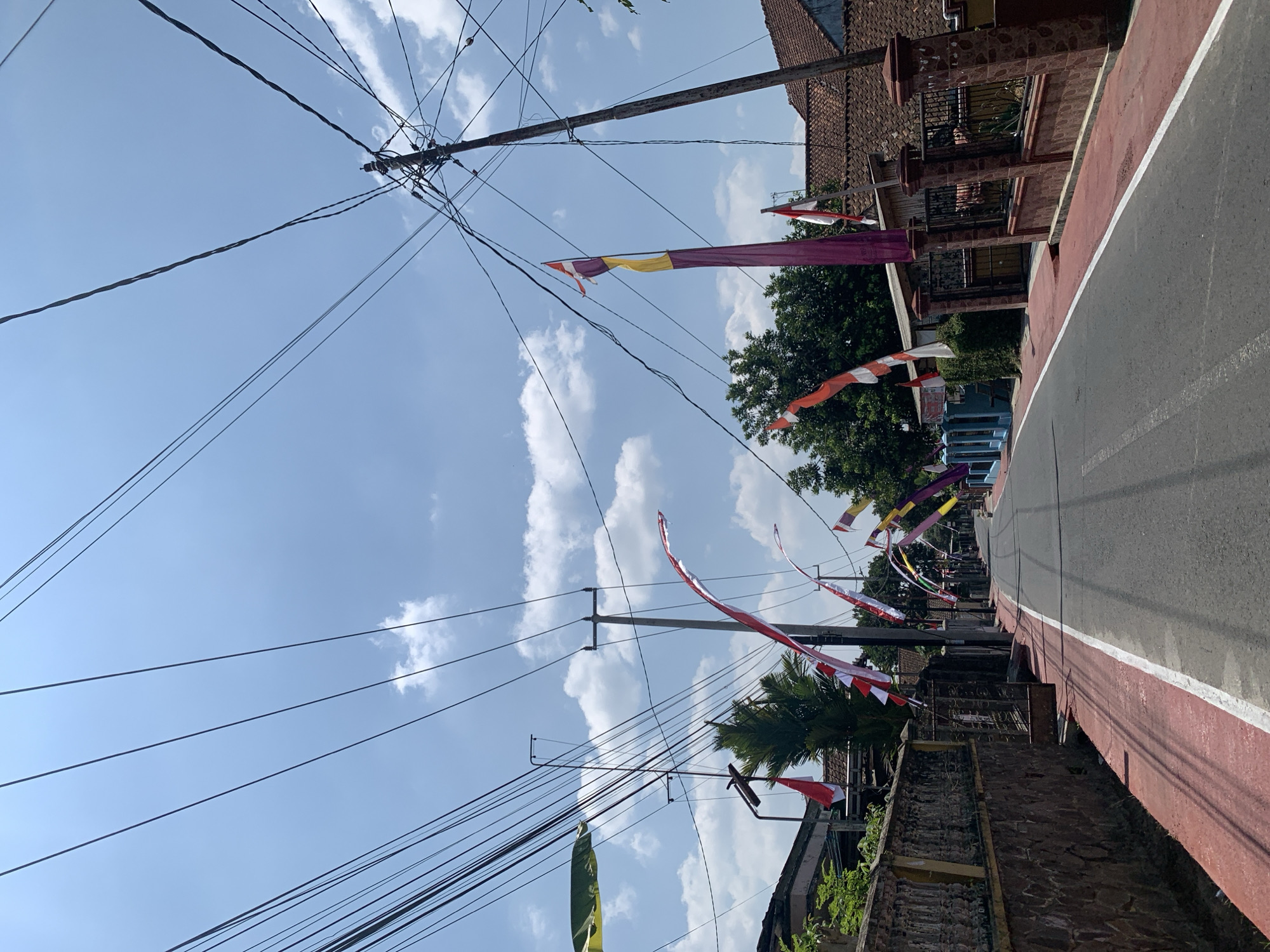 The image depicts a street in the city of Ciamis, Indonesia, adorned with numerous Indonesian flags, likely for a national celebration. The street is paved and has a red painted sidewalk on the left side with a white line marking the edge of the road. The flags, predominantly red and white, are hung along the street on poles and wires, creating a festive atmosphere. Electrical wires crisscross the sky, supported by utility poles. Houses with tiled roofs line the street, with some visible fences and walls. The sky is blue with scattered white clouds, indicating a clear day. No people are visible in the image. The overall scene suggests a public street that has been decorated for a patriotic event.