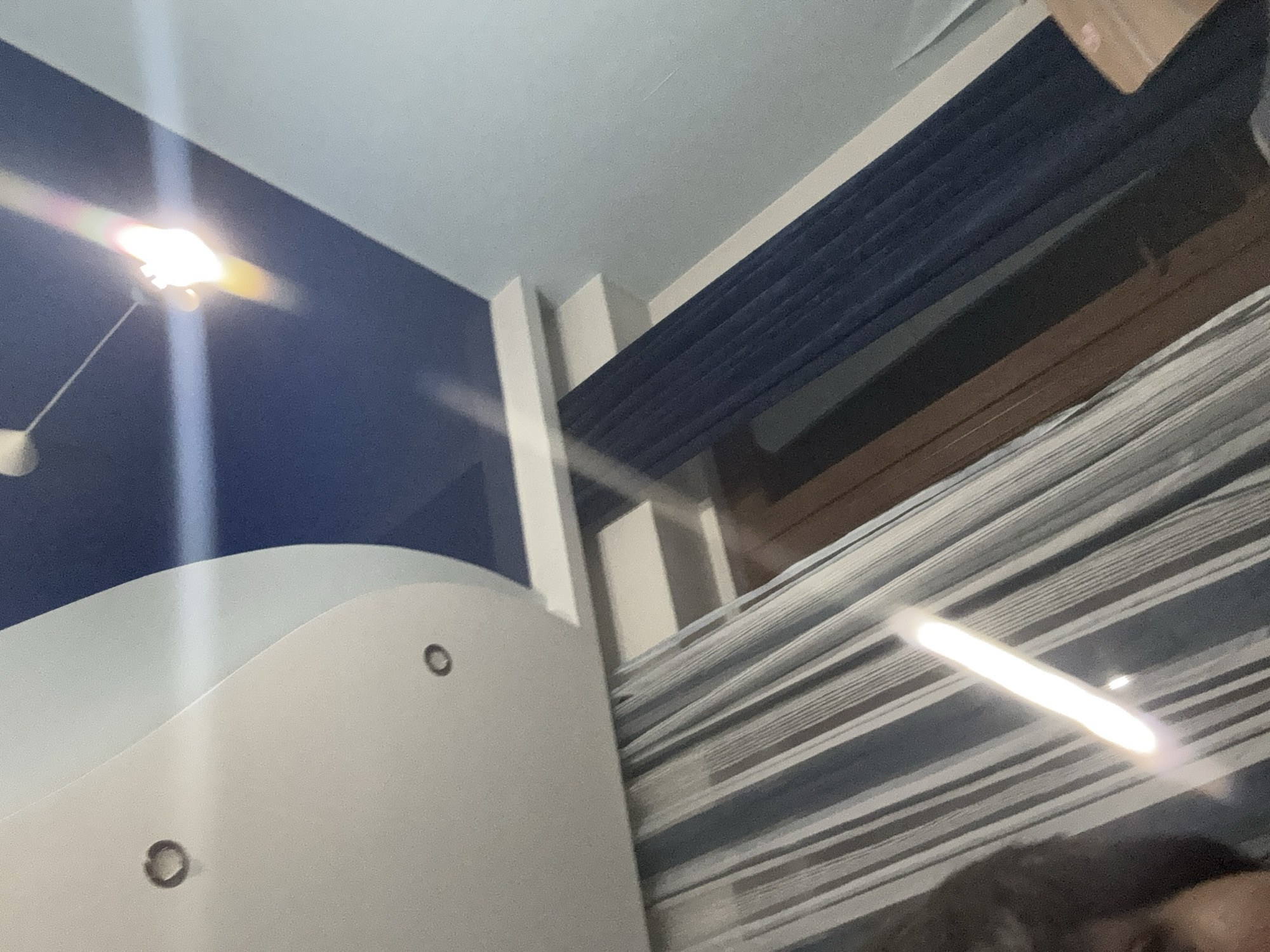 An indoor scene shows a ceiling with a dark blue section and a white curved section with recessed lighting fixtures. A pendant light with a bright bulb hangs from the blue section, casting a strong glare. Below the ceiling, striped curtains and dark blue curtains are visible. Part of a person's face is visible in the lower left corner, reflected in the curtains. A wooden door frame is also present. The overall impression is of a room with a modern design.