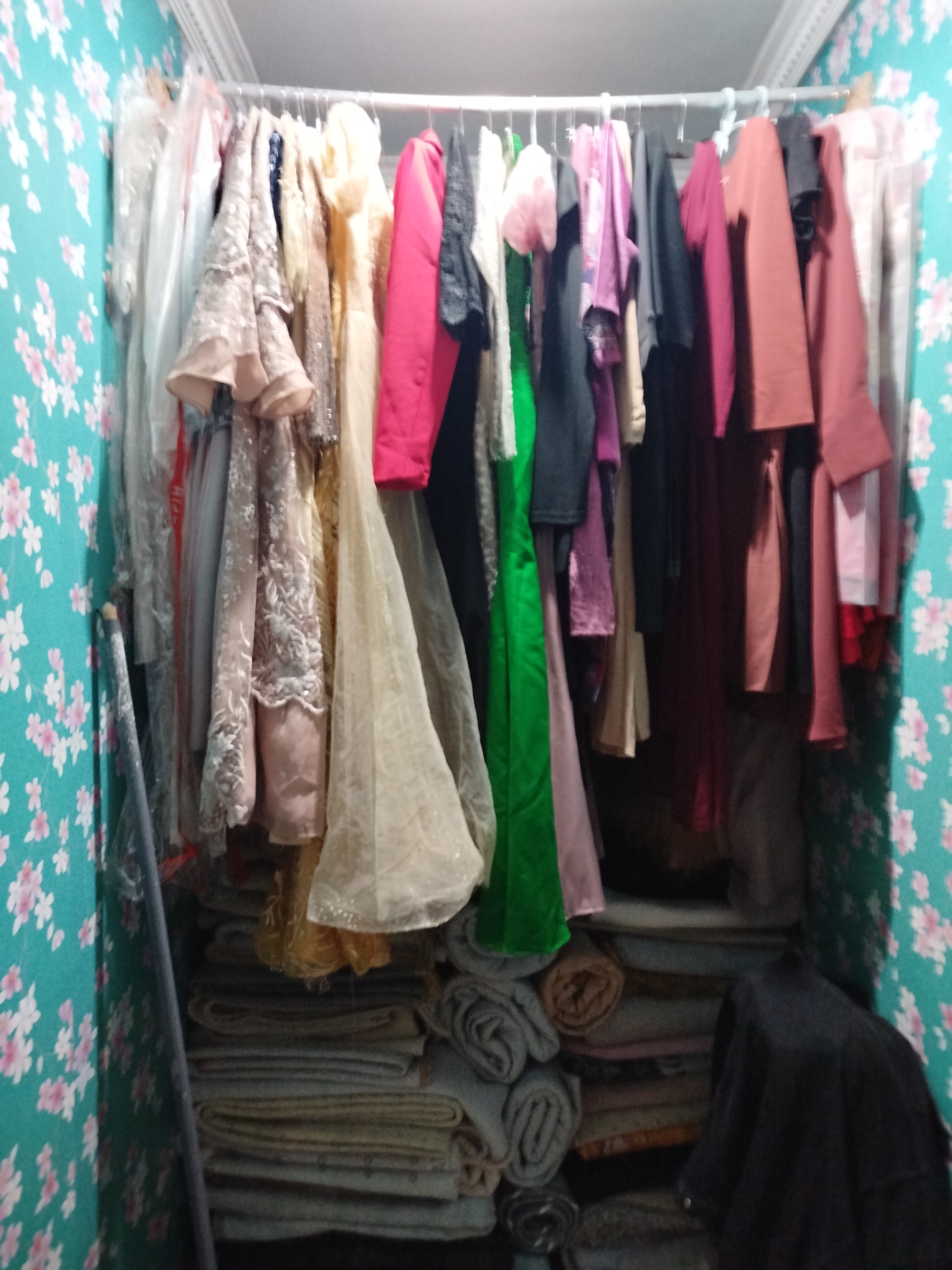 The image shows a closet filled with clothes hanging on a rack and folded on shelves. The walls are covered with teal wallpaper featuring a floral pattern of pink and white flowers. The closet is illuminated by overhead lighting, casting shadows. On the rack, there are various garments, including dresses and blouses, in a range of colors such as pink, red, beige, gold, and black. Some of the clothes appear to be formal wear, with embellishments and flowing fabrics. The shelves below are stacked with folded textiles, possibly blankets or more clothing. To the left, a long, slender object, likely a broom or mop handle, leans against the wall. To the right, a dark-colored piece of fabric, possibly a garment bag or a draped cloth, is visible. The scene appears to be in a residential setting, with the floral wallpaper suggesting a decorative choice for the room. The city of Bandungrejo, Indonesia, is mentioned as the location, but no specific landmarks or contextual elements within the image directly indicate this.