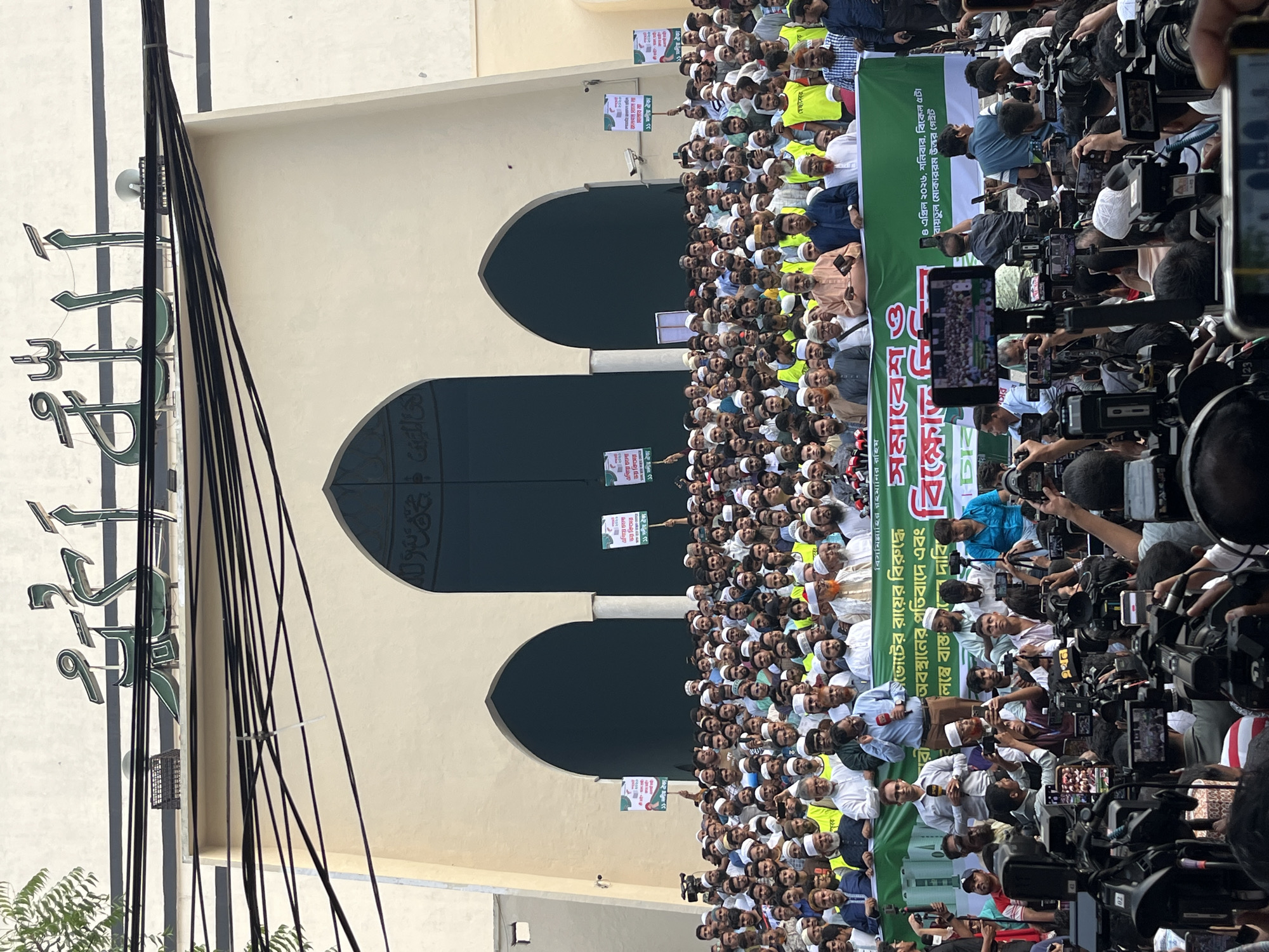 The image depicts a large gathering of people in front of a building with three arched entrances. Many of the people are wearing white caps, and some are holding signs and banners. A group of journalists with cameras and microphones is gathered in the foreground. The building appears to be a mosque, based on the architectural style and the Arabic script on the front. Based on the text on the signs, it is likely a protest or demonstration related to a political issue. The signs refer to an "11-party alliance" and mention "Baitul Mukarram North Gate," which is a location in Dhaka, Bangladesh.