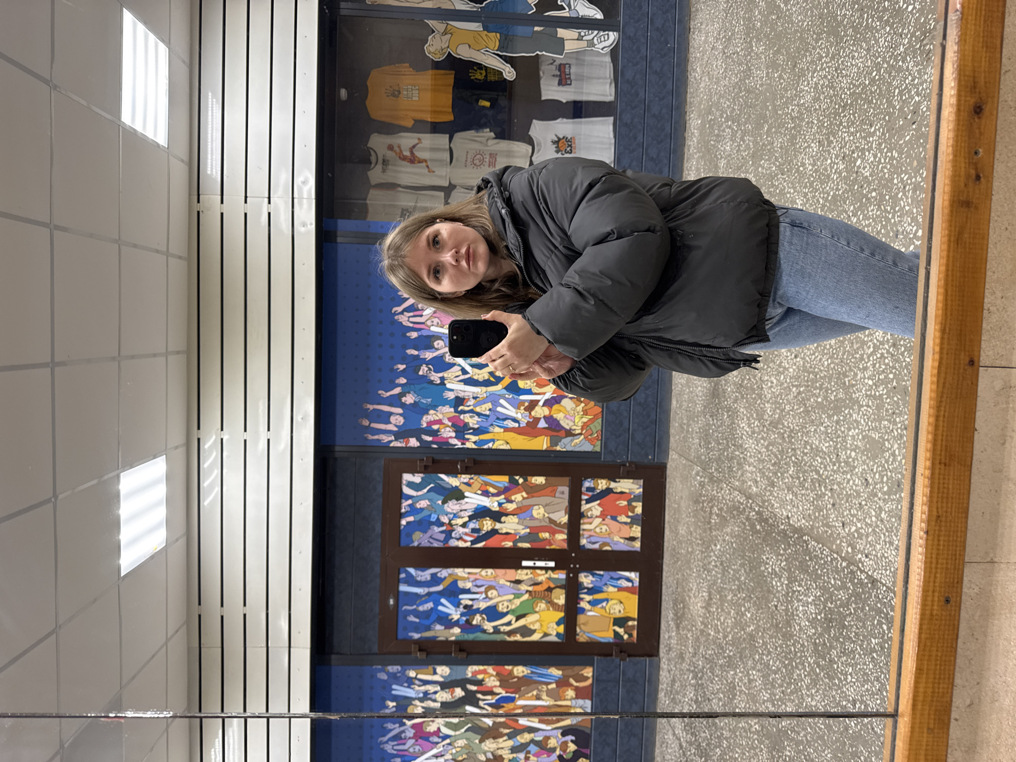 A person is reflected in a mirror, holding a cell phone. The reflection shows a view of a building interior. The building appears to be a retail store or school. A display case with clothing is visible. A mural of many figures is also visible. The floor is gray with a speckled pattern. The wall is white with linear indentations. The city of Perm, Russia is not mentioned.