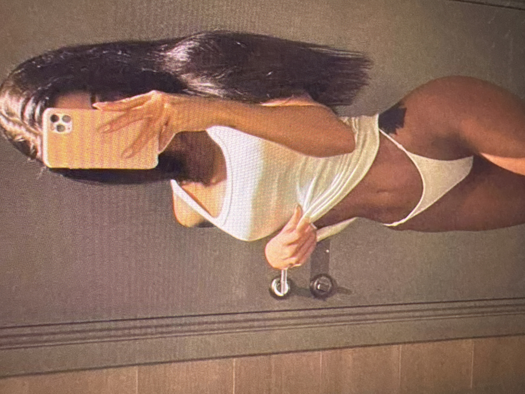 The image shows a person taking a selfie in what appears to be a bathroom. The person is wearing a white tank top and white underwear. They are holding a light orange smartphone to take the picture. The background consists of a door and part of a wall. There is a doorknob and a keyhole visible on the door. There is also a tattoo visible on the person's hip. The image is low resolution and grainy. The location in the city of Castelo Branco, Portugal cannot be determined from the image.