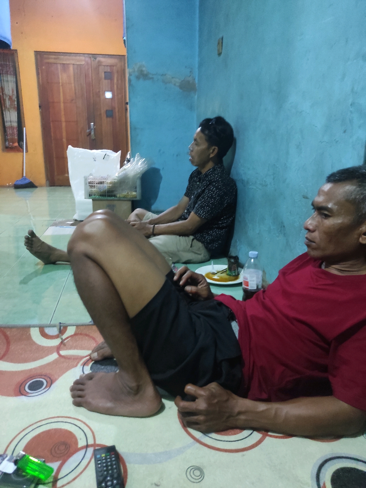 The image depicts an indoor scene, possibly a home, with two men present. The man in the foreground, wearing a red t-shirt and black shorts, is lying on his side on a patterned rug. His right arm is extended, and his hand rests near his thigh. His left leg is bent, with his foot visible. The man in the background, wearing a dark patterned shirt and light-colored pants, is sitting with his back against a blue wall, facing to the left. A white plastic bag and a cage containing items are placed behind him. Other objects visible include a bottle of water, a plate with food, a remote control, and a green lighter. The floor appears to be tiled in a light green color. A wooden door is visible on the left side of the image, along with a broom leaning against the wall. The overall environment suggests a casual, domestic setting. The location is identified as Laksanamekar, Indonesia.