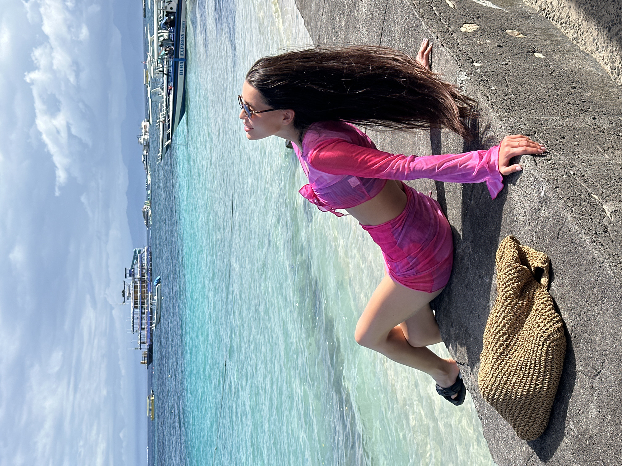 A woman with long, dark hair and sunglasses is leaning on a concrete wall. She wears a long-sleeved pink crop top and a matching pink skirt. Her hair is blown back. A woven beach bag sits beside her. Behind the wall is the sea. Boats are visible on the water under a cloudy sky. This location is in Nusa Penida, Indonesia.