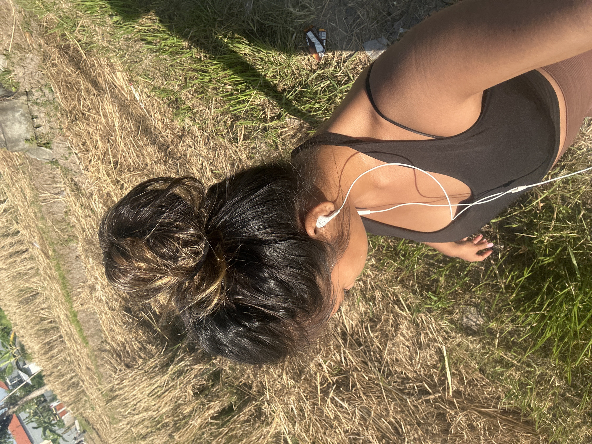 A person with dark hair styled in a messy bun, wearing a black tank top and white earphones, is standing in a dry, grassy field with some green grass and shadows from a bright sun. The location appears to be a rice paddy field after harvest in Sukawati, Indonesia. In the background, there are buildings and trees, suggesting a rural or semi-rural environment. There are also some discarded items on the ground, including what appears to be snack wrappers. The person is looking down and to their left.