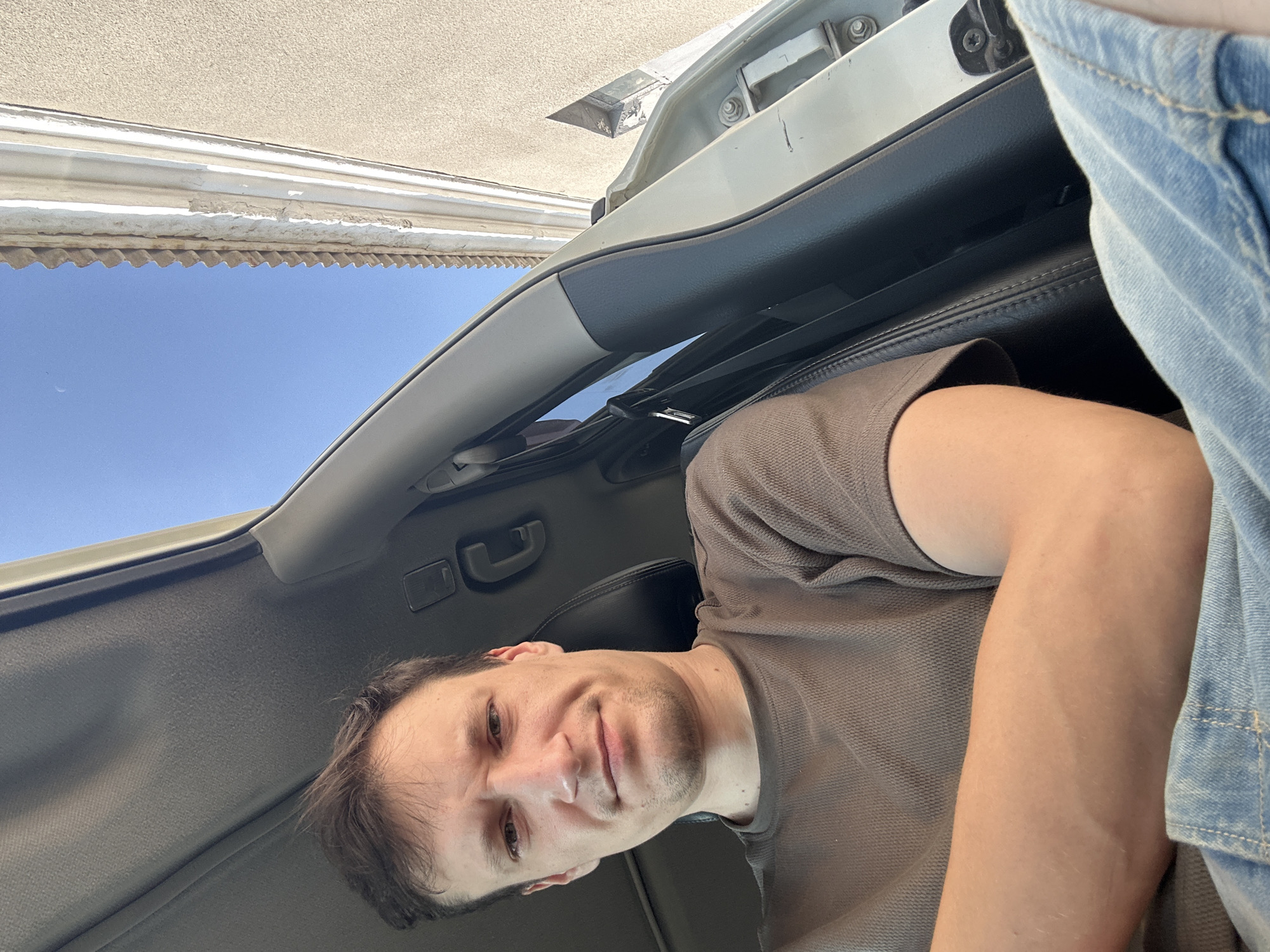 A man is sitting in the driver's seat of a car, looking at the camera. The car's sunroof is open, revealing a clear blue sky and a building with a corrugated roof. The man is wearing a brown t-shirt and denim shorts. The interior of the car appears to be dark leather. The man's left arm is resting on his lap, and his right arm is bent at the elbow. The image was taken outdoors in Shatava, Ukraine.