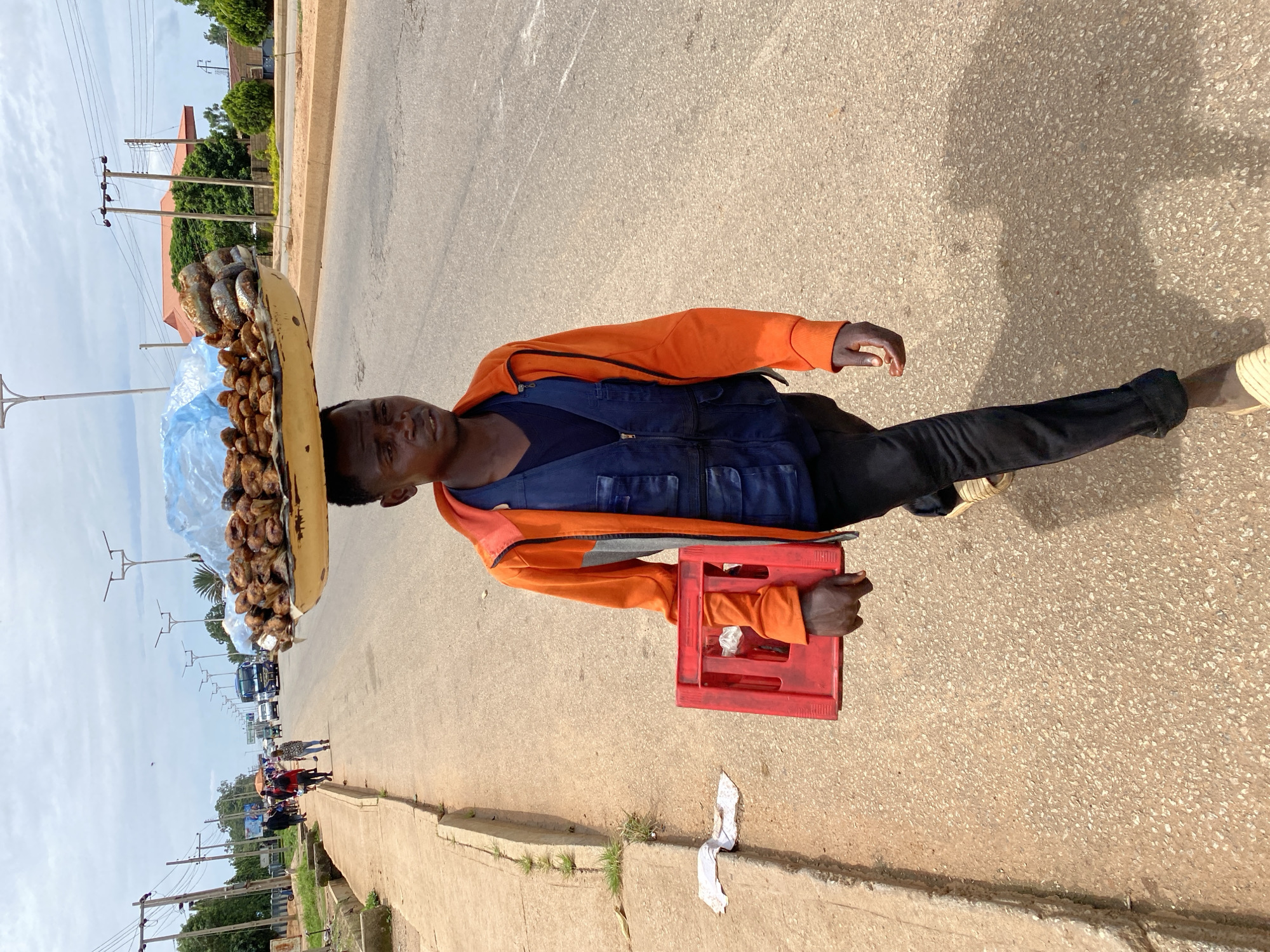 A young Black male walks on a paved road in Bukuru, Nigeria, carrying a large platter of fried fish balanced on his head. He wears an orange hooded jacket over a dark blue vest and black pants. In his right arm, he holds a red plastic crate containing white plastic bags. The road is flanked by a concrete sidewalk with some sparse vegetation and litter. In the background, various vehicles, including a bus, are visible on the road, along with utility poles and buildings. The sky is cloudy.