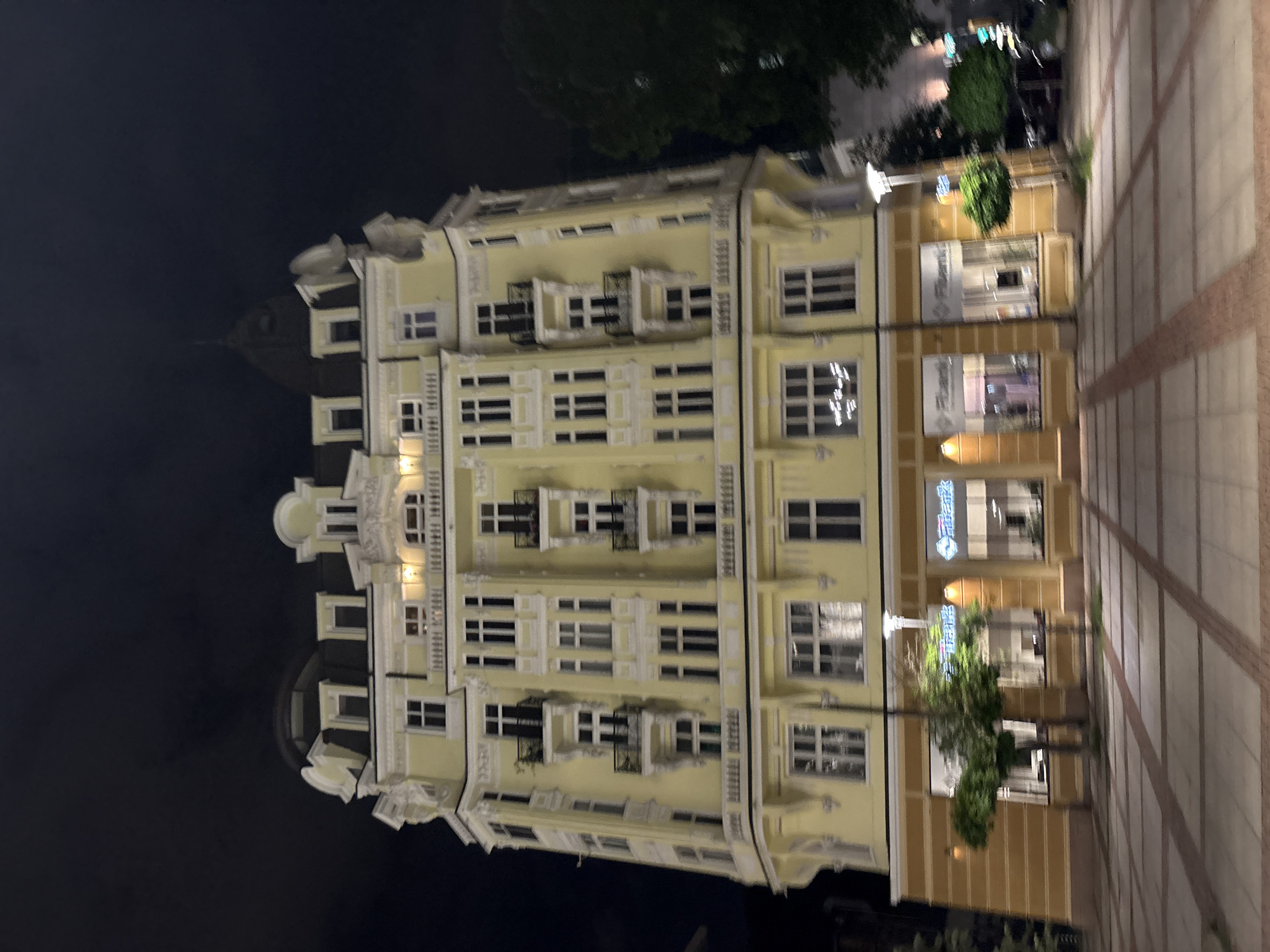 The image shows a multi-story yellow building with ornate architectural details, including balconies and decorative trim, in Sofia, Bulgaria. The building appears to be divided into sections, with the right side featuring storefronts with "Fibank" signage. A paved plaza with trees and lamp posts is visible in front of the building. The sky is dark, suggesting it is nighttime. There are no visible people in the frame.