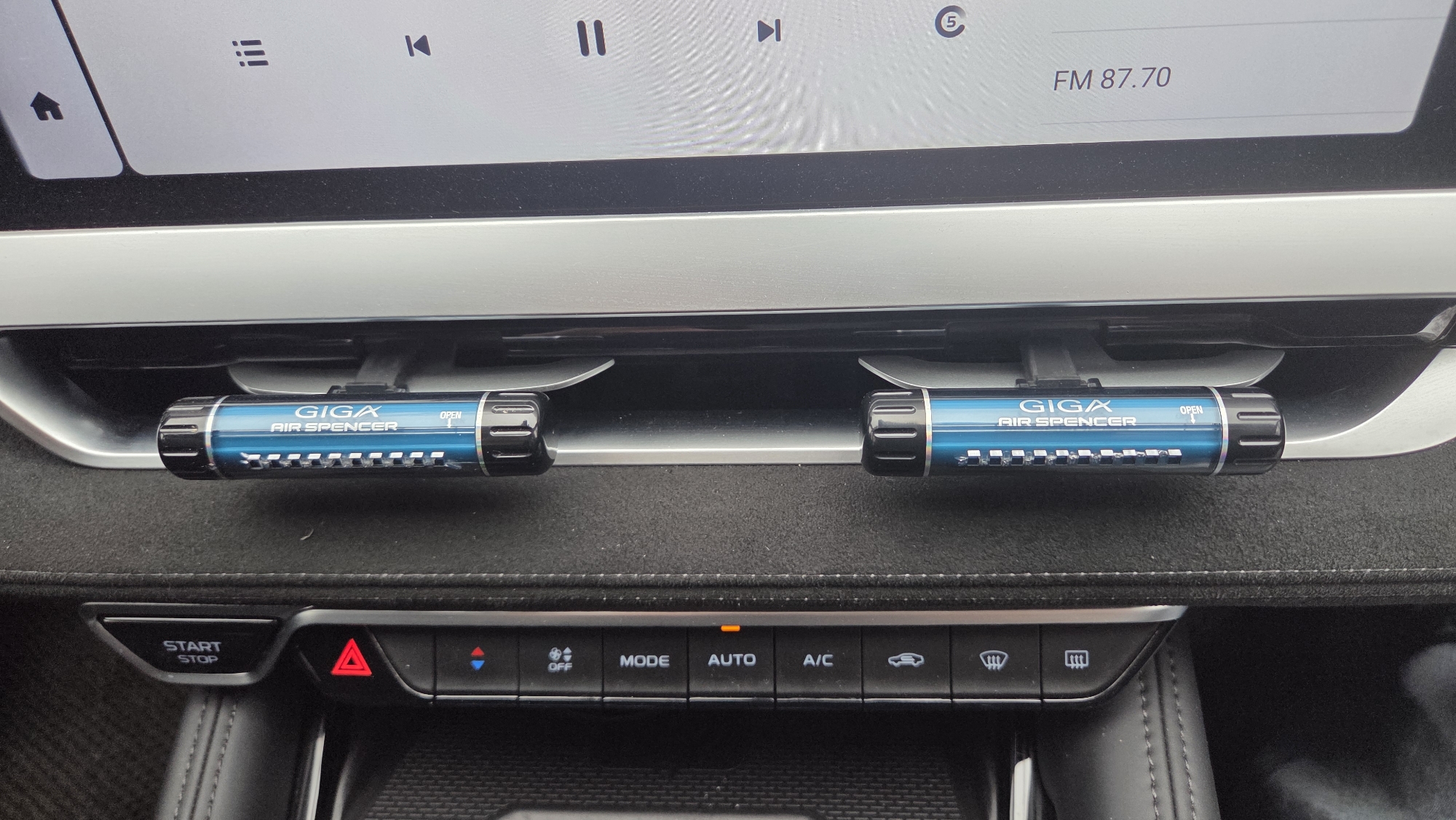 The image shows the interior of a vehicle, specifically the dashboard area below a large infotainment screen. Two cylindrical air fresheners, labeled "GIGA AIR SPENCER" in blue with black ends, are mounted on the air vents beneath a silver-colored horizontal trim piece. The dashboard is covered in a dark material, possibly suede or a similar soft textile. Below the air fresheners, a row of buttons controls various vehicle functions, including hazard lights, climate control (OFF, MODE, AUTO, A/C), defrosters, and other settings. To the left of the buttons is a "START STOP" button. The infotainment screen above displays various icons, including a home button, a list icon, media control buttons (pause, skip), and a radio frequency of FM 87.70. The overall environment suggests the interior of a modern vehicle. There is no indication of location in Chaikovsky, Russia.