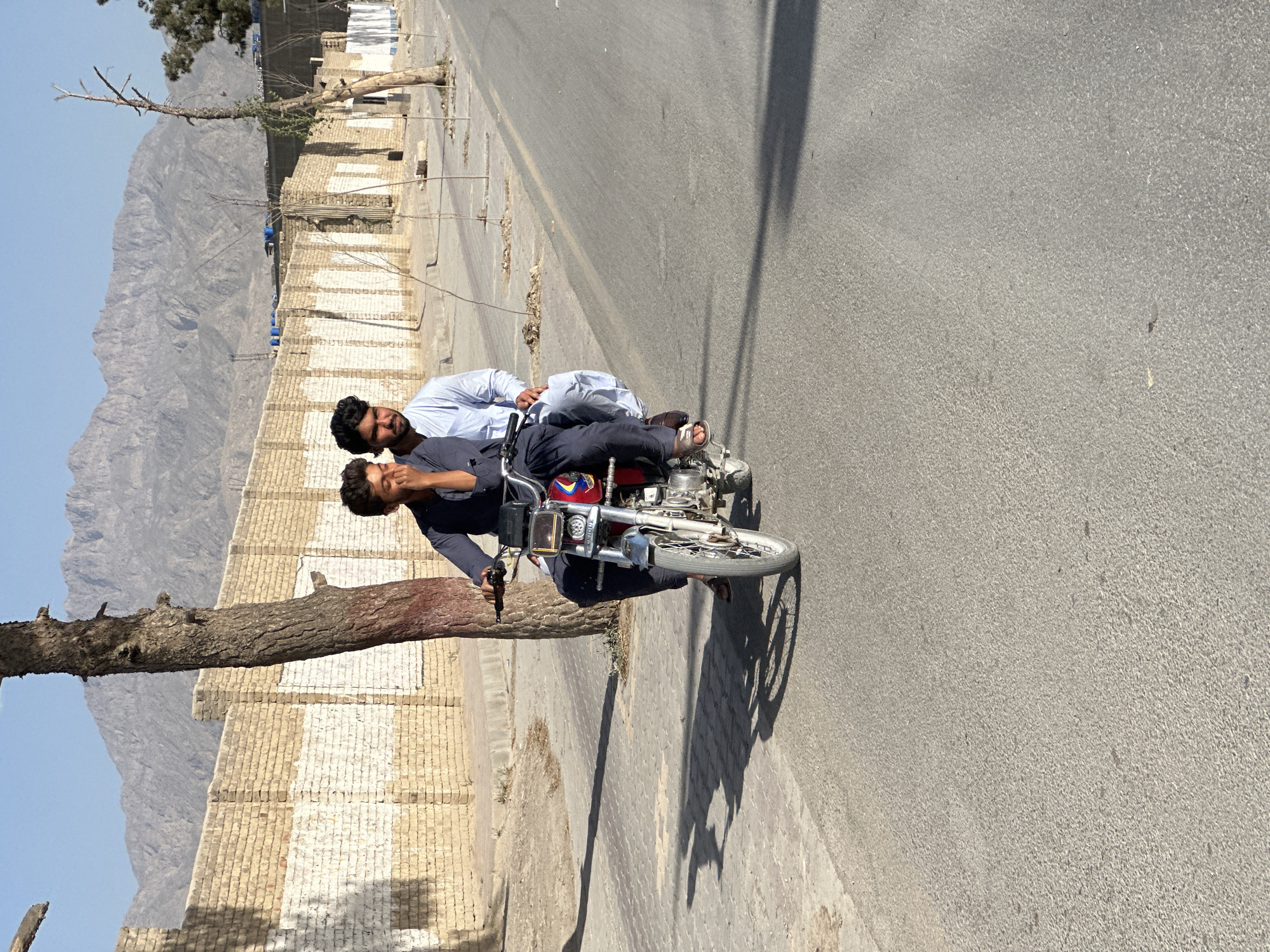 The image shows two men on a motorcycle on a road. One man is driving and the other is riding behind him. The road appears to be within a city but the location is not identified. There are buildings on one side of the road and a mountain in the distance.