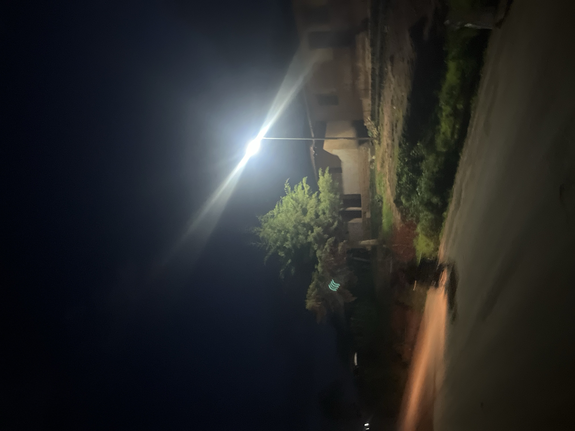 This is a nighttime, low-light photograph taken in Gembu, Nigeria. The scene depicts a street with a single lamppost illuminating a portion of the area. The lamppost is tall and emits a bright, white light that creates a strong lens flare diagonally across the upper portion of the image. To the right of the lamppost, there is a building constructed from what appears to be mud or earth-toned materials, with visible windows. Adjacent to the lamppost and in front of the building is a mature, green tree. The foreground shows a paved road, with some of the surface appearing wet or reflecting light, suggesting it may have rained or there are puddles. The headlights of a moving vehicle, possibly a motorcycle, are visible on the road, casting an orange glow and creating a motion blur, indicating movement. The surrounding environment appears to be somewhat rural or on the outskirts of a settlement, with vegetation visible along the side of the road and in front of the building. The sky is dark, characteristic of nighttime.