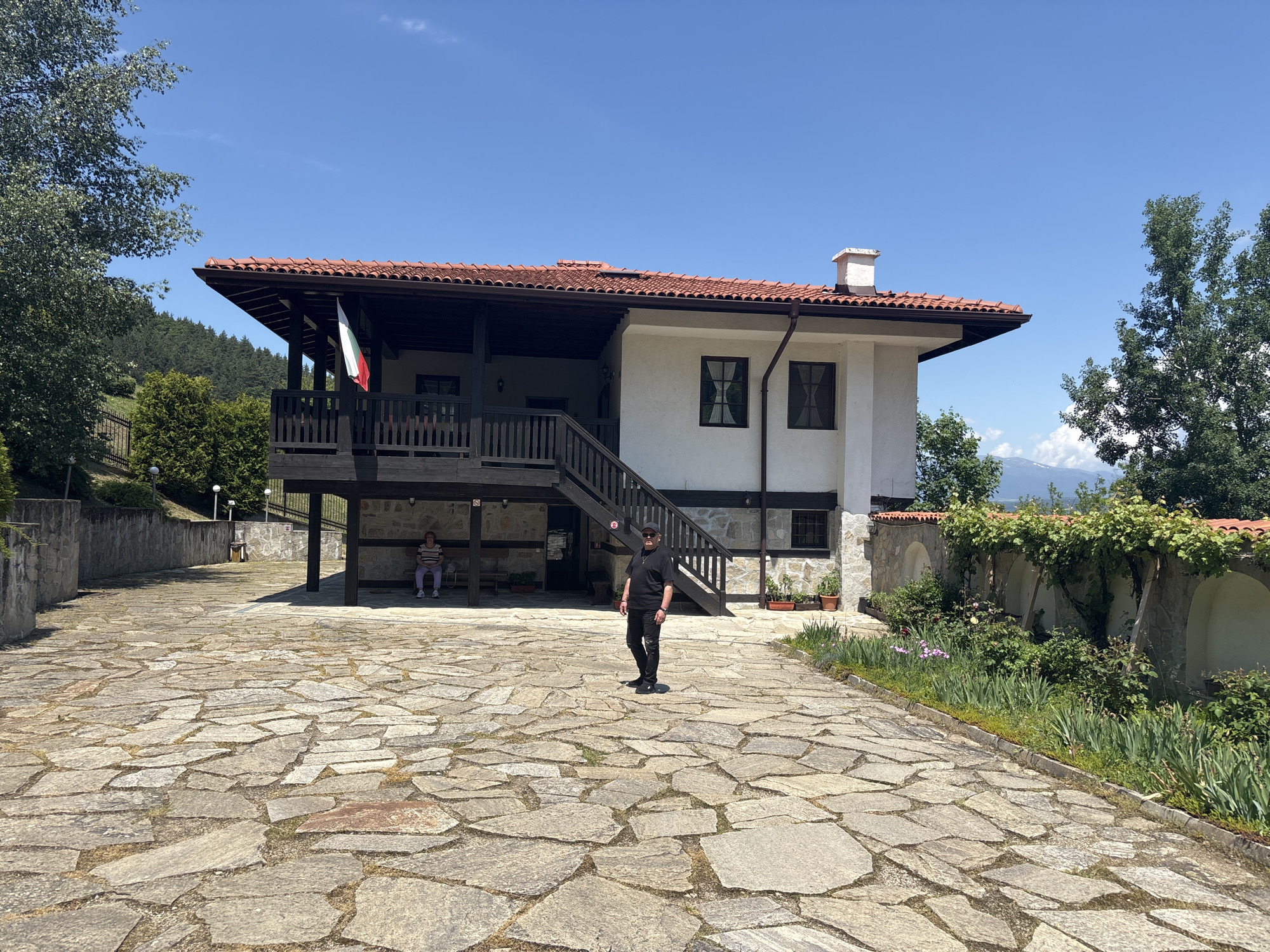 The image depicts an outdoor scene in Belchin, Bulgaria, on a clear, sunny day. A two-story building with a red-tiled roof and white facade on the upper level occupies the center-right. A wooden balcony with railings extends from the left side of the upper floor, with a Bulgarian flag (white, green, red horizontal stripes) hanging vertically from it. A wooden staircase leads from the ground to this balcony. Windows with dark frames are visible on the building's facade. A white chimney is present on the right side of the roof.
The foreground features a large courtyard paved with irregularly shaped stone slabs, with sparse grass growing between them. A man stands centrally in the courtyard, facing slightly left of the camera. He is dressed in a black t-shirt, black trousers, black shoes, a black baseball cap, and dark sunglasses. His posture is upright, with his hands at his sides.
On the left side of the building, beneath the balcony, a woman is seated on a wooden bench in a shaded area. She wears a light-colored top, light-colored trousers, and white shoes, and appears to be holding a small object in her hands while facing away from the camera's direct view.
To the far left, a large deciduous tree with green foliage is visible. The background on the left features a hillside covered with evergreen trees and other vegetation, partially obscured by a dark metal fence. Along the right side of the courtyard, a low stone wall with several arched recesses is covered in green vines and plants, with various flowers and potted plants arranged in front of it. Distant mountains, possibly with remnants of snow or light clouds, are visible on the horizon to the far right. The sky is bright blue with minimal scattered clouds.