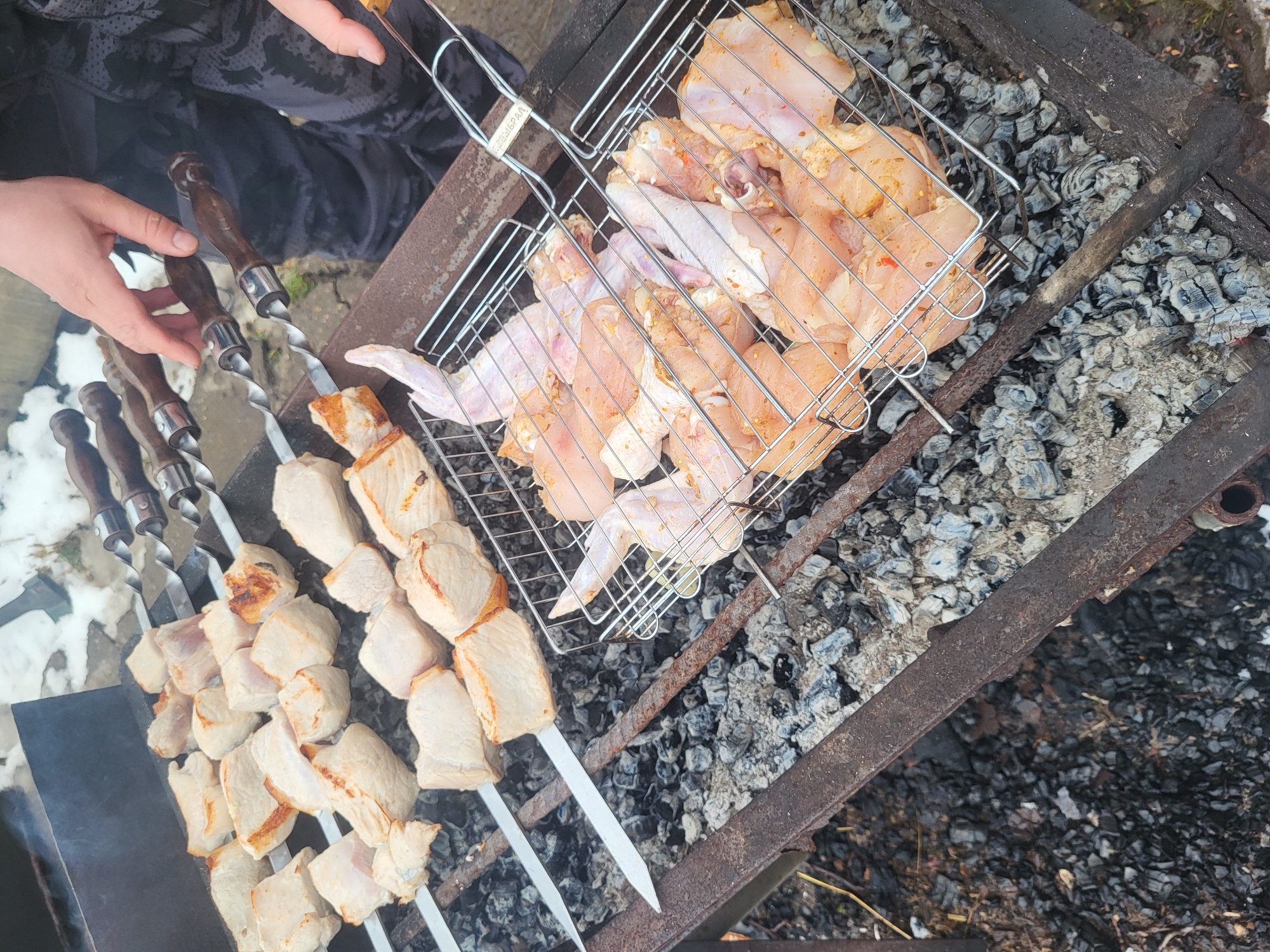 The image depicts a grill with food being cooked. On the grill are skewers of meat and a metal basket containing raw chicken pieces. The grill is placed over hot coals. A person is holding a skewer. The location is likely outdoors. The city is Зипуново, Russia.