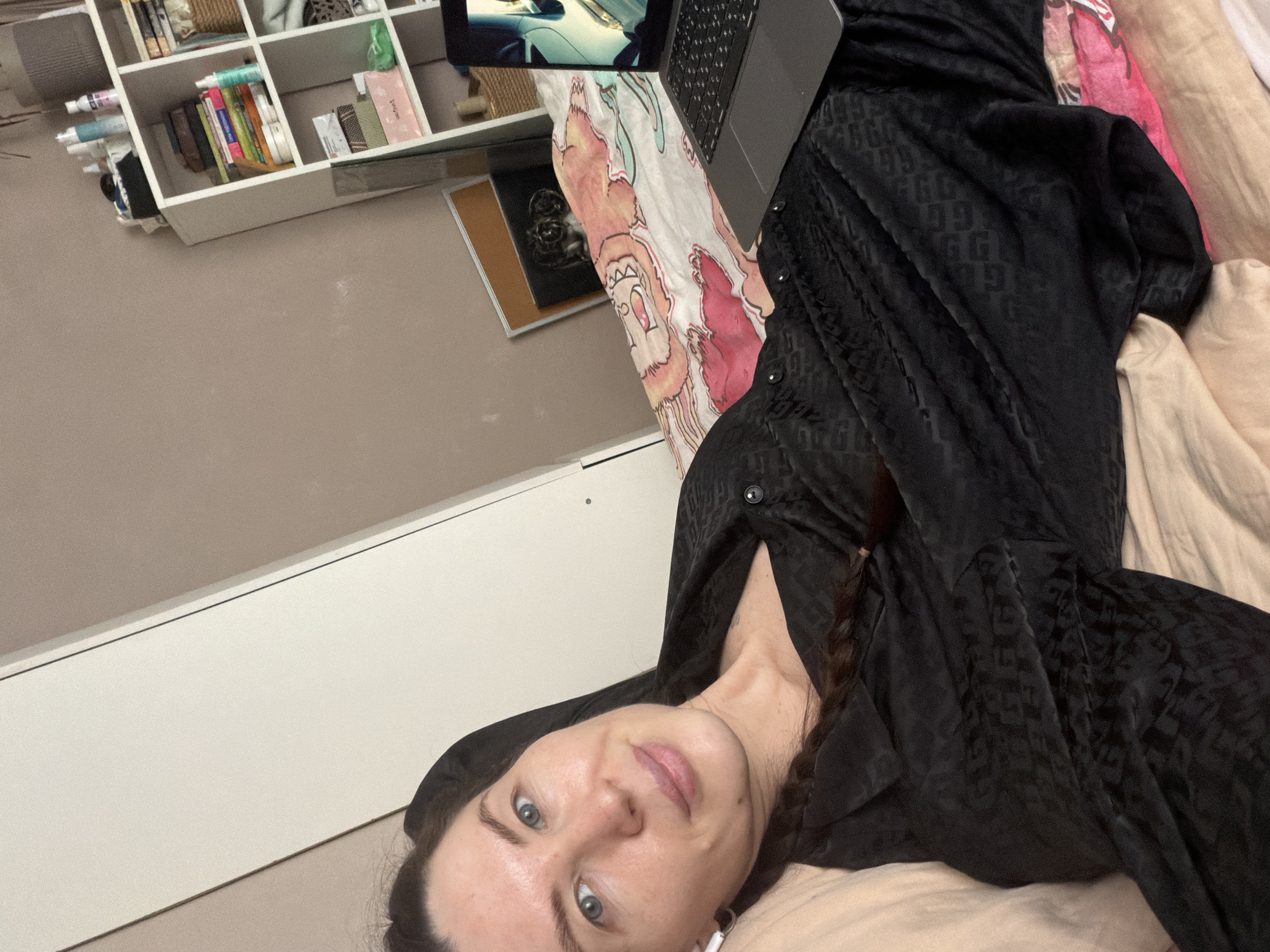 An adult female, estimated to be between 30 and 40 years old, is depicted in a semi-recumbent position on a bed in a domestic indoor setting. She has fair skin, light blue eyes, and dark hair, part of which is visible as a braid on her left side. She wears a dark, possibly black, patterned collared shirt or pajama top and a white wireless earbud in her left ear. Her gaze is directed towards the camera, and she exhibits a slight smile.
To her right, a silver-grey laptop is open on the bed, displaying illuminated content that appears to be a stylized image or video. The bed is covered with a multi-colored duvet featuring cartoonish figures in pink, orange, and white. Beyond the laptop and duvet, a pair of animal eyes is visible, suggesting the presence of a small pet peeking over the duvet.
The background includes a light brown/taupe wall. To the left, a light-colored wardrobe or closet with two vertical doors is partially visible. To the right, a white open-shelf bookcase is present, stocked with various items including bottles, books, baskets, and boxes. A framed item leans against the wall below the bookcase. The specific location within the city of Perm, Russia, cannot be determined from the visual evidence presented in the image.