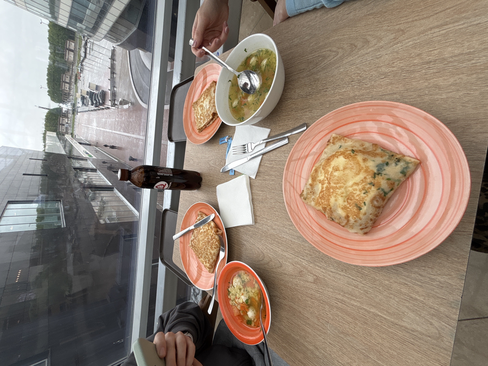 The photograph captures a meal in what appears to be a modern restaurant or café, with a view through a large window that overlooks a cityscape likely within Saint Petersburg, Russia, judging by the architectural style and the presence of Cyrillic characters on a beverage bottle. The window shows a wet urban environment, suggesting it has recently rained.
Several plates of food are arranged on a wooden table. Two of the plates contain what appear to be folded pancakes, possibly crepes, with one displaying green herbs. Two additional plates contain soup with ingredients such as vegetables and what seem to be small meatballs or dumplings, with a spoon in one of the bowls. Cutlery is neatly arranged on napkins, alongside the plates. A bottle of dark-colored liquid, perhaps a soft drink, is also visible near the window.
Parts of individuals are visible. A hand is reaching for the soup with a spoon, and another person is visible with a cell phone. The setting suggests a casual dining experience, where the patrons are enjoying their meal while observing the city outside.