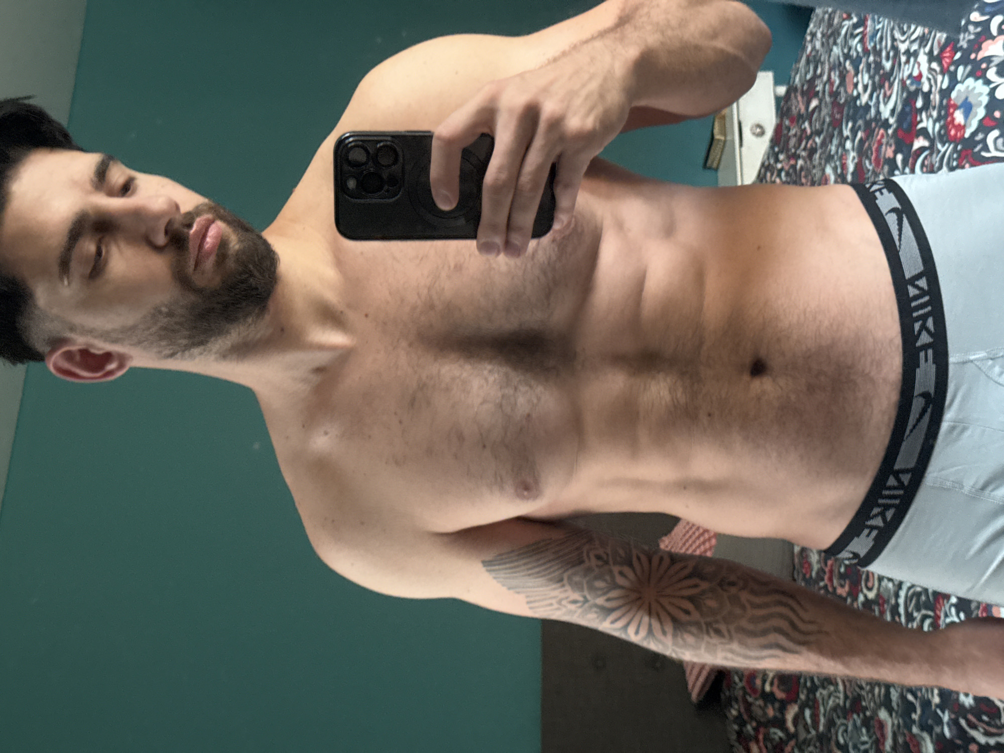 The image is a mirror selfie depicting a man, presumably in a residence in Sofia, Bulgaria. The man, seen from the chest up, is shirtless, revealing a fit physique with visible abdominal muscles and some chest hair. He has dark hair and a beard, and his eyes appear partially closed, suggesting a relaxed or contemplative mood. His right hand holds a black smartphone with multiple camera lenses, used to capture the image. A distinctive tattoo featuring a mandala-like pattern and wavy designs covers his left forearm. He is wearing light grey boxer briefs with a black waistband that prominently displays the word "NIKE" in white capital letters.
The background, visible as a reflection, includes a teal-colored wall on the left side of the frame. To the right, part of a bed with a dark, intricately patterned duvet or comforter is visible. The bedding features a colorful design with red, white, blue, and green floral or abstract motifs. The overall setting appears to be an indoor space, likely a bedroom or possibly a bathroom, under ambient indoor lighting. The primary activity is the man taking his own picture in a mirror.