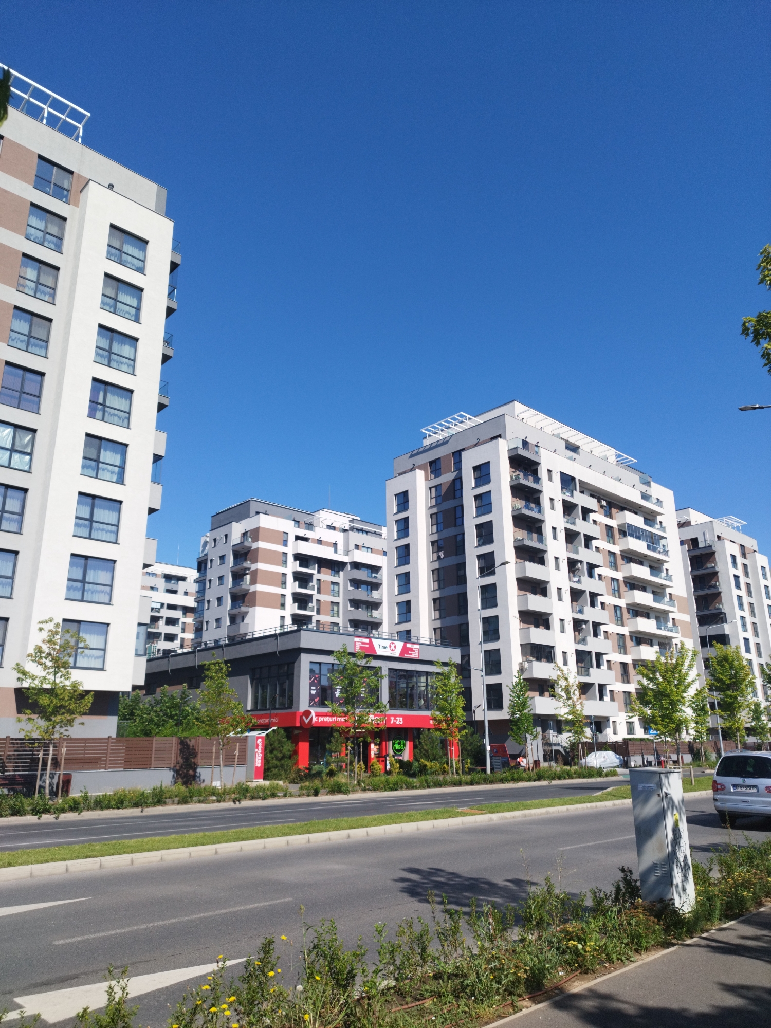 The image shows a street scene in Bucharest, Romania, with modern apartment buildings lining the road. The buildings are contemporary in design, featuring large windows, balconies, and a mix of white, grey, and brown facades. A street with a double lane of traffic runs horizontally across the foreground, bordered by a grassy median and sidewalks. On the right side of the road, a silver car is visible. Along the base of the buildings, there are trees and shrubs. A red-and-white storefront with the logo "Food" is visible on one of the buildings, suggesting a commercial or retail space. The sky is clear and blue, indicating a sunny day. The overall impression is one of urban development and a modern residential area.