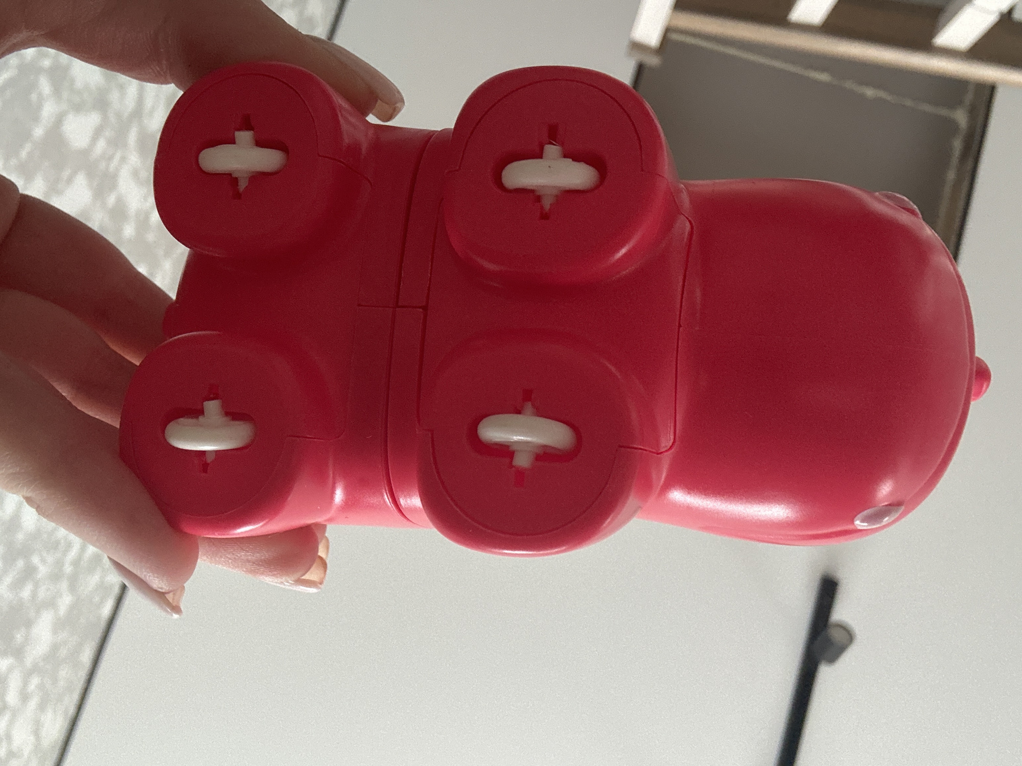 A red plastic hippo toy, seen from below, is being held in a person's hand. The toy has four circular feet with white cross-shaped plastic elements protruding from them. The hippo's body appears to be composed of multiple sections. The location is unspecified, but the prompt mentions Chaikovsky, Russia. The environment is generic and includes a white surface.