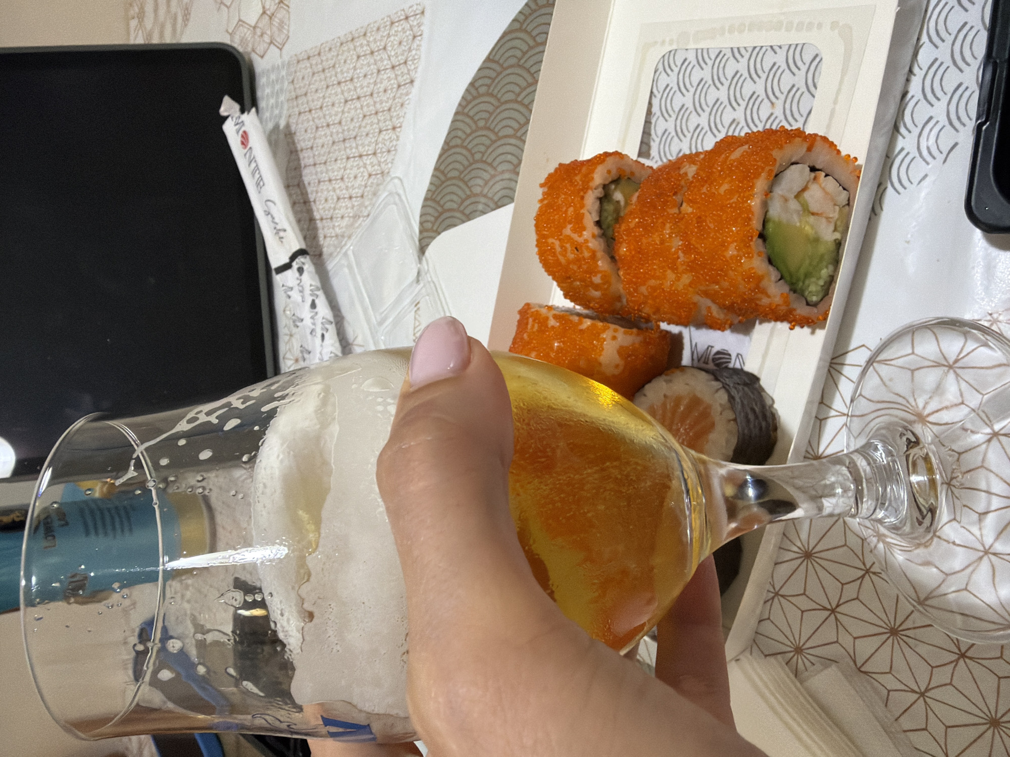 A close-up shot captures a person's hand holding a glass of beer with foam, and a tray of sushi rolls. The beer is golden yellow and has a frothy head. The sushi rolls are topped with orange roe and appear to be California rolls with avocado and shrimp. The background includes chopsticks, a laptop, and a patterned tablecloth. The scene is indoors, likely in a home or restaurant setting. There is no specific indication of the location being in the city of Budva, Montenegro.