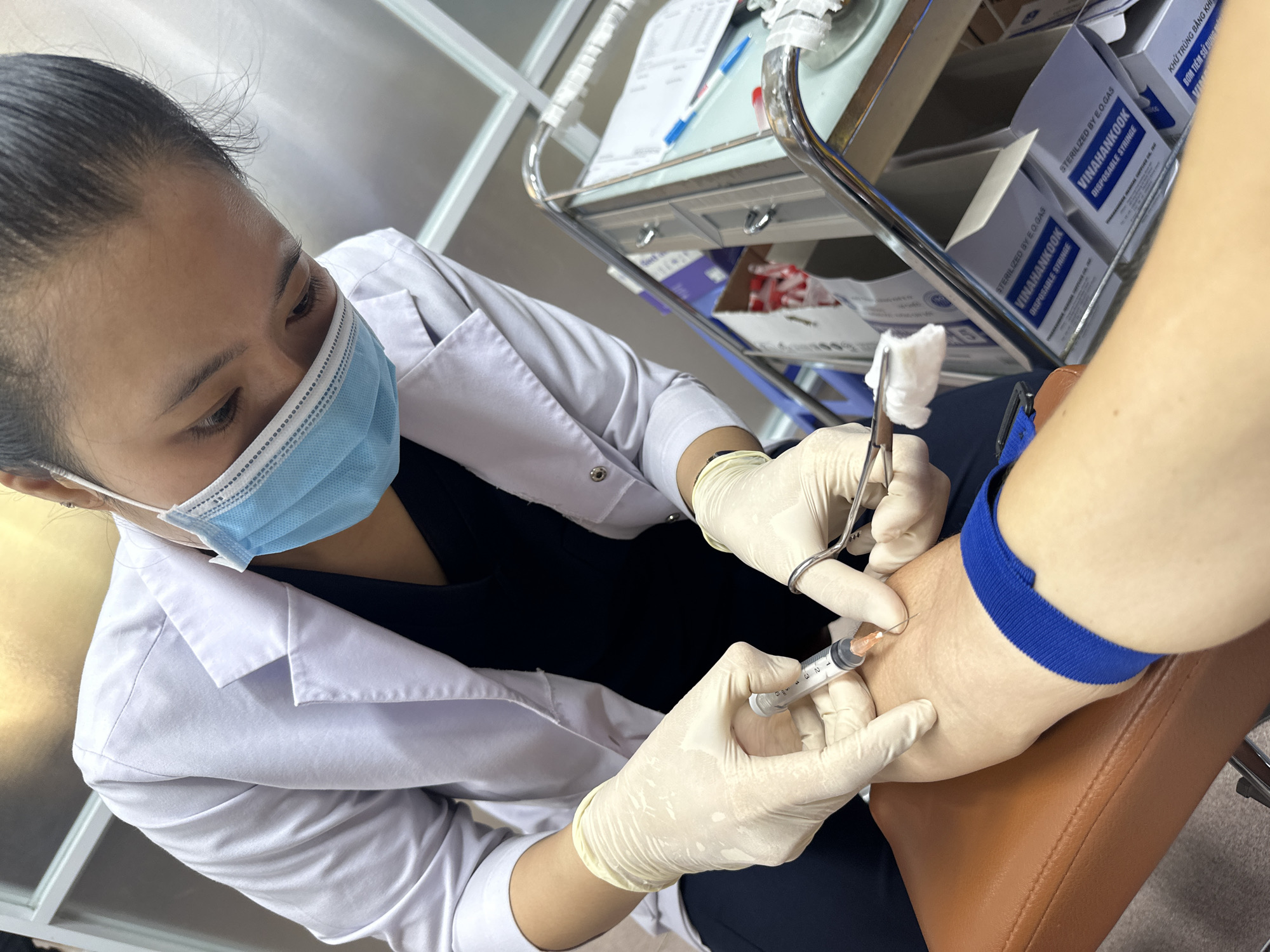 A person wearing a white lab coat, light-colored gloves, and a light blue surgical mask is in the process of drawing blood from a person's arm. The person on the left is holding a syringe and a pair of scissors with gauze. The person on the right has their arm resting on a brown cushion, with a blue tourniquet applied to their upper arm. The scene appears to be a medical setting, possibly a clinic or hospital, with medical equipment and supplies visible in the background, including boxes labeled "VINAHANKOOK DISPOSABLE SYRINGE." The overall environment is clean and sterile. The specific location in Nha Trang Ward, Vietnam is not evident from the image.
