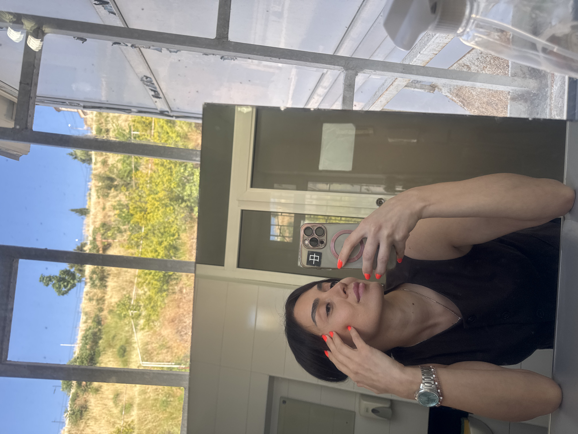 The image shows a woman taking a selfie in a bathroom with a mirror reflecting the outdoors and the bathroom interior. The woman has dark hair and is wearing a dark sleeveless top. Her fingernails are painted bright orange, and she is wearing a silver watch with a blue face on her left wrist. She is touching her face with her right hand, and holding a smartphone in her left hand.
The bathroom appears to have tiled walls and a large mirror. Behind the woman, a doorway or window leads to an outdoor scene. The outdoor scene features a grassy, hilly landscape with some trees and buildings in the distance, under a clear blue sky. The overall impression is of a sunny day. The context of Tbilisi, Georgia is inferred due to the specific architectural style of the window frames and the hilly terrain, which is characteristic of the city. The presence of a clear blue sky suggests it's likely daytime, and the vegetation indicates a warmer season. The metallic structure visible on the right side of the image could be part of the building's exterior or a window frame.