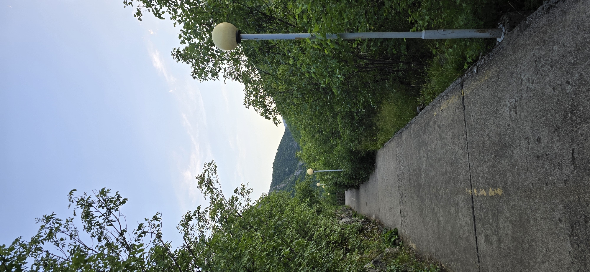 The image shows a paved pathway lined with lush green trees and vegetation, leading towards a mountainous landscape under a clear sky. Several street lamps with yellow globes are spaced along the right side of the path. The pathway itself is made of concrete with some yellow markings. The scene is natural and appears to be a park or a scenic route. Based on visual cues, this location is likely the path leading to the Vratsata Gorge or the Vratzata Rocks, popular natural attractions in Vratsa, Bulgaria, known for their hiking trails and scenic beauty.