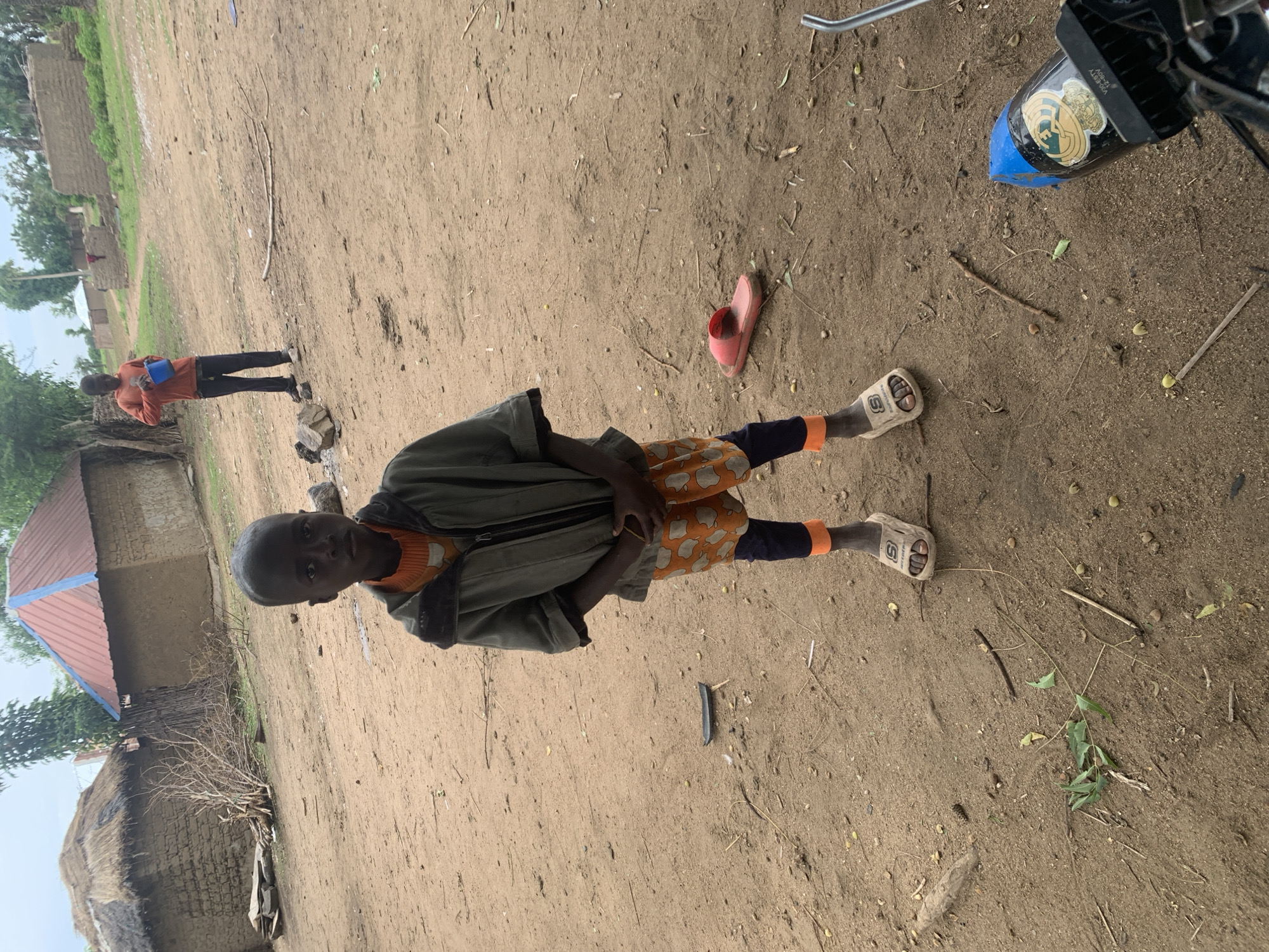 A young boy stands in the foreground of the image, looking directly at the camera. He is wearing a grey hooded jacket over an orange shirt and orange shorts with a repeating elephant pattern. His legs are covered by dark blue leggings that end in bright orange cuffs. He wears a single white sandal on his left foot. His right foot is bare. The ground is dry, sandy earth with scattered leaves and twigs.
In the background, two traditional huts are visible. The hut on the left has a thatched roof, while the hut on the right has a red metal roof with blue trim. A second, older boy or young man is standing further back, on the right side of the frame, holding a blue bucket and looking to his left. Beyond the huts, more buildings and trees can be seen under a cloudy sky. The scene appears to be a rural village in Dull A, Nigeria.
