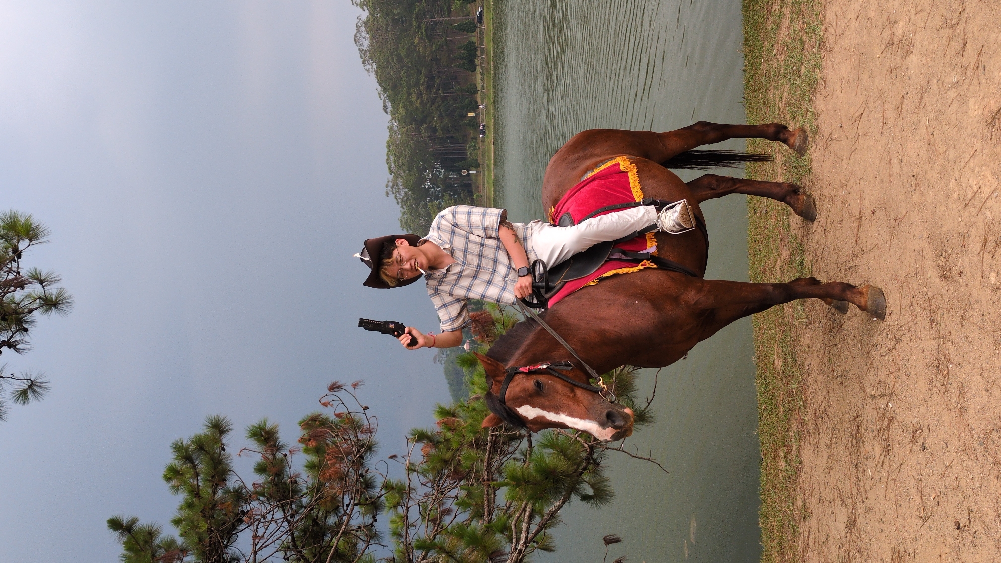 A person wearing a brown cowboy hat, eyeglasses, a white and blue plaid shirt, and white pants is riding a brown horse near a body of water, possibly a lake, in Da Lat, Vietnam. The horse has a red and gold saddle blanket. The person is holding a black pistol in their right hand. Trees are visible in the background and foreground, and there's a strip of land or path beside the water. The sky is overcast. The location is likely near a tourist attraction or park within Da Lat.