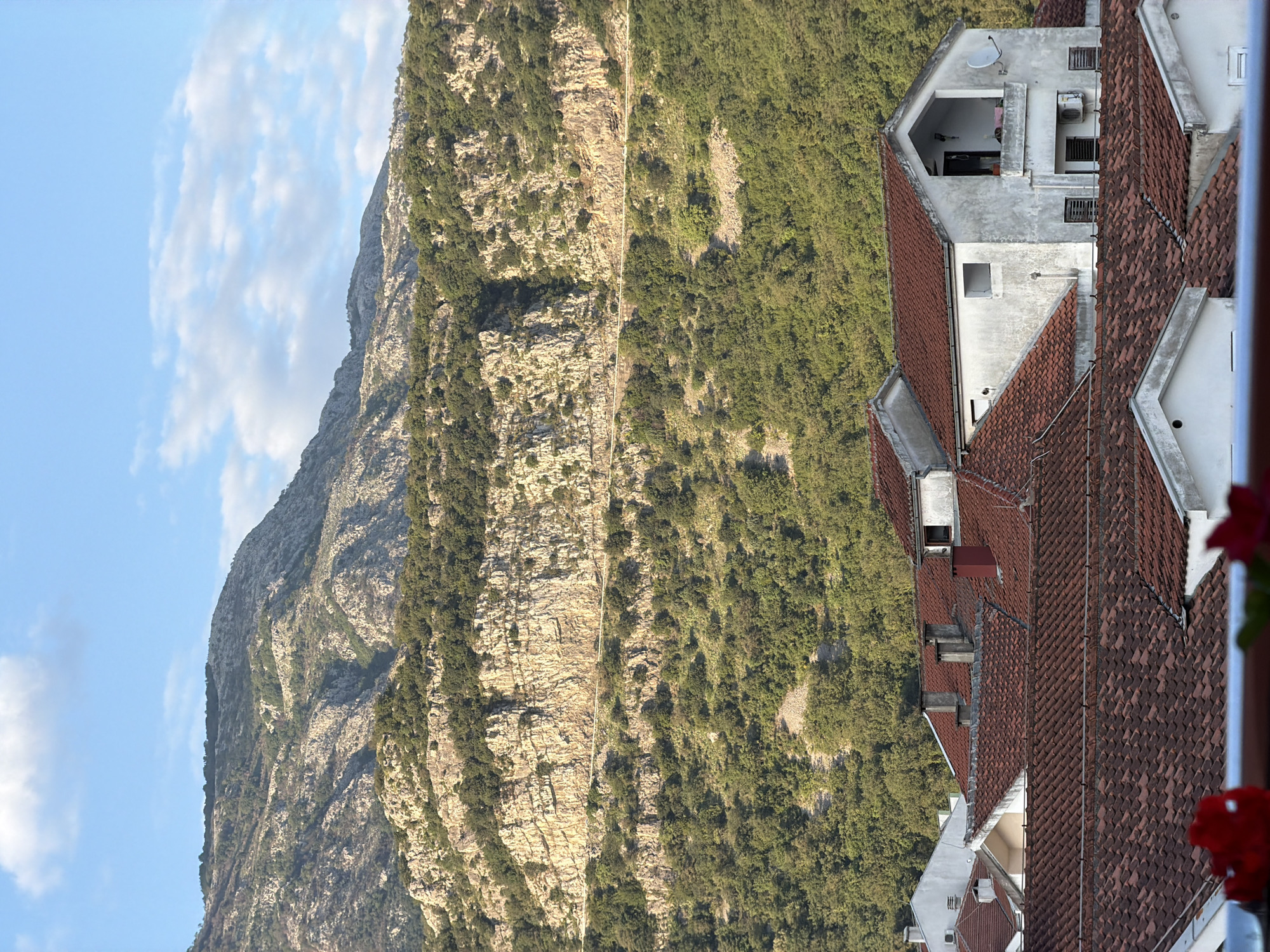 The image shows a view of a mountain landscape in Budva, Montenegro, observed from an elevated position, possibly a building or balcony. In the foreground, the red tiled roofs of buildings are visible. The architecture includes elements of light-colored plaster and more modern additions. The backdrop features a rugged mountain with a mix of exposed rock faces and verdant vegetation. The sky is partly cloudy, with blue patches interspersed among the white clouds. The image is taken in daylight.