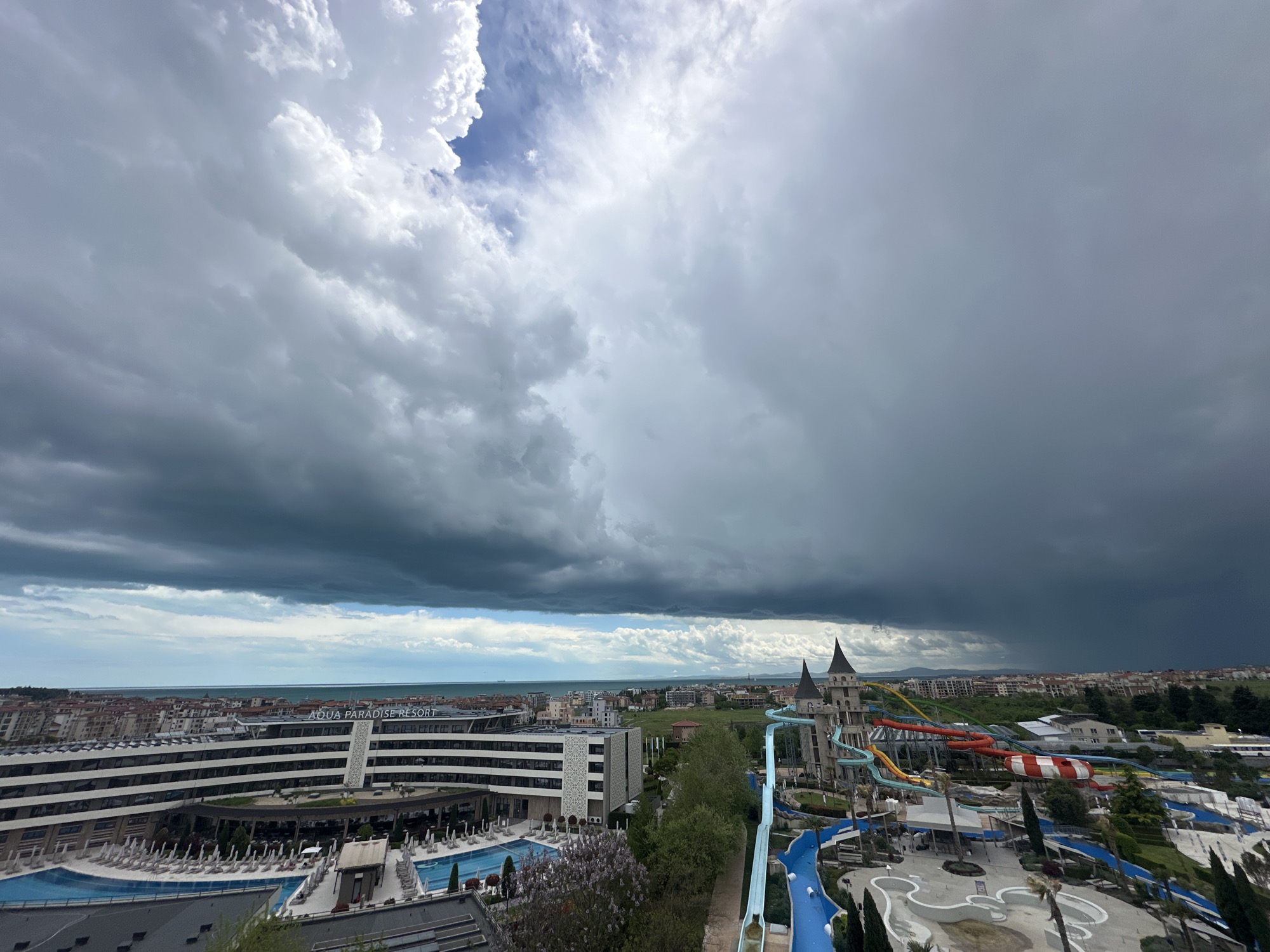 The image shows a panoramic view of Ravda, Bulgaria, dominated by a gathering storm. In the foreground is the Aqua Paradise Resort, featuring a large hotel complex with white and grey architecture, pools with lounge chairs, and a water park with colourful slides and castle-like structures. The water park appears deserted, likely due to the impending storm. Beyond the resort, the urban landscape of Ravda is visible, characterized by buildings of varying heights and colours. In the distance, the Black Sea is discernible, with a visible horizon line. The sky is heavily overcast with dark grey storm clouds looming over the city, contrasting with a small patch of blue sky and white clouds.