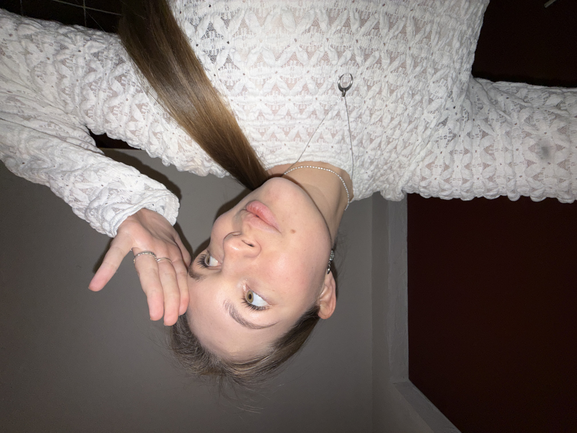 The image features a young woman captured in an indoor setting. She is positioned upside down in the original photo, but described here as if upright. She has light brown, long hair, parted to one side, and fair skin. Her eyes are light-colored, possibly green or hazel, and she is looking upwards and slightly to her left. Her expression appears thoughtful or contemplative.
She is wearing a white, long-sleeved top made of a textured fabric with an intricate, open-knit or lace-like pattern. The sleeves also feature this detailed pattern. A delicate silver-toned necklace is visible around her neck, with a distinctive ring-shaped pendant resting on her chest. Small silver earrings are visible in her ears, and she wears several rings on the fingers of her right hand, which is raised to her temple or forehead area, with fingers slightly spread.
The background is simple, featuring a dark grey wall to her left and a darker, possibly deep red or brown, surface to her right. A vertical white strip, likely a door frame or architectural element, is visible on the far right. The lighting is soft and even, suggesting indoor artificial light rather than bright daylight. There is no visible text in the image. The location context provided is Belgrade, Serbia, though no specific visual cues confirm this within the image itself.