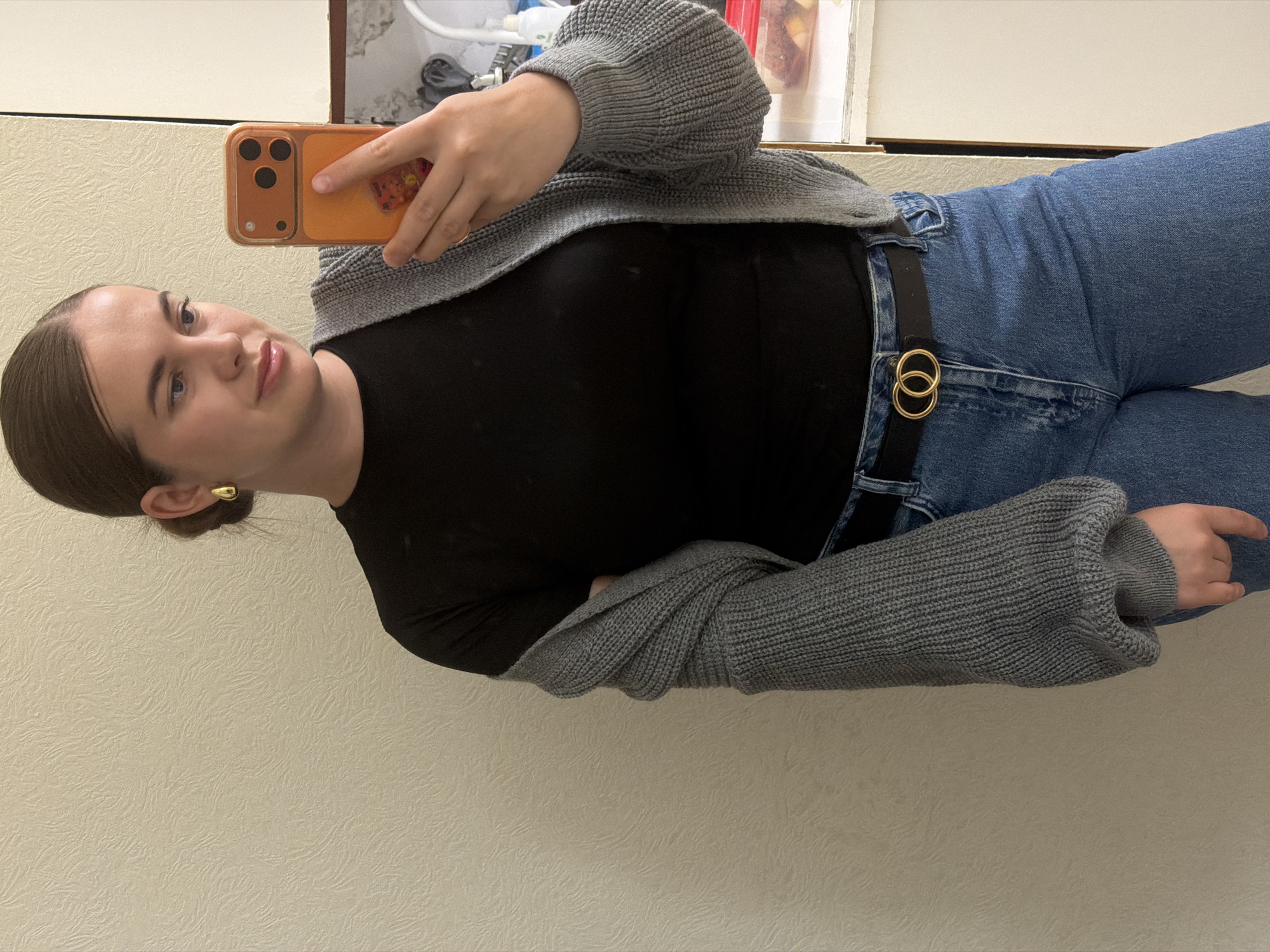 A young woman is depicted in a mirror selfie, with the image rotated 90 degrees clockwise. She is presumed to be in Sochi, Russia.
**Subjects:** The primary subject is a young woman, likely in her 20s, with dark hair pulled back into a neat bun. She has light skin, light eyes, and a calm expression with a subtle hint of a smile. She is wearing a black long-sleeved top, a grey chunky knit cardigan with voluminous sleeves, and blue denim jeans. A black belt with a distinctive golden double-ring buckle is visible at her waist. She wears a small, polished gold hoop earring in her visible ear.
**Objects:** She is holding an orange smartphone, possibly an iPhone due to its triple-camera setup, in her right hand to capture the selfie. Her clothing items—the black top, grey cardigan, blue jeans, and black belt with a gold buckle—are clearly visible.
**Scene/Setting:** The scene is indoors, likely in a room with a light-colored, textured wall forming the main background. In the upper part of the image, there's a glimpse of a reflective surface, possibly a mirror or glossy countertop, and some indistinct objects on a shelf or surface, suggesting a bathroom or dressing area.
**Activity or event taking place:** The woman is actively taking a selfie using her smartphone, holding it up towards the reflective surface to capture her image.
**Notable details:** The lighting is soft and even, typical of indoor settings. The woman's posture is relaxed. The overall mood is casual. The image is oriented horizontally, requiring a rotation to view upright.
**Visible text:** No discernible text is visible anywhere in the image.
