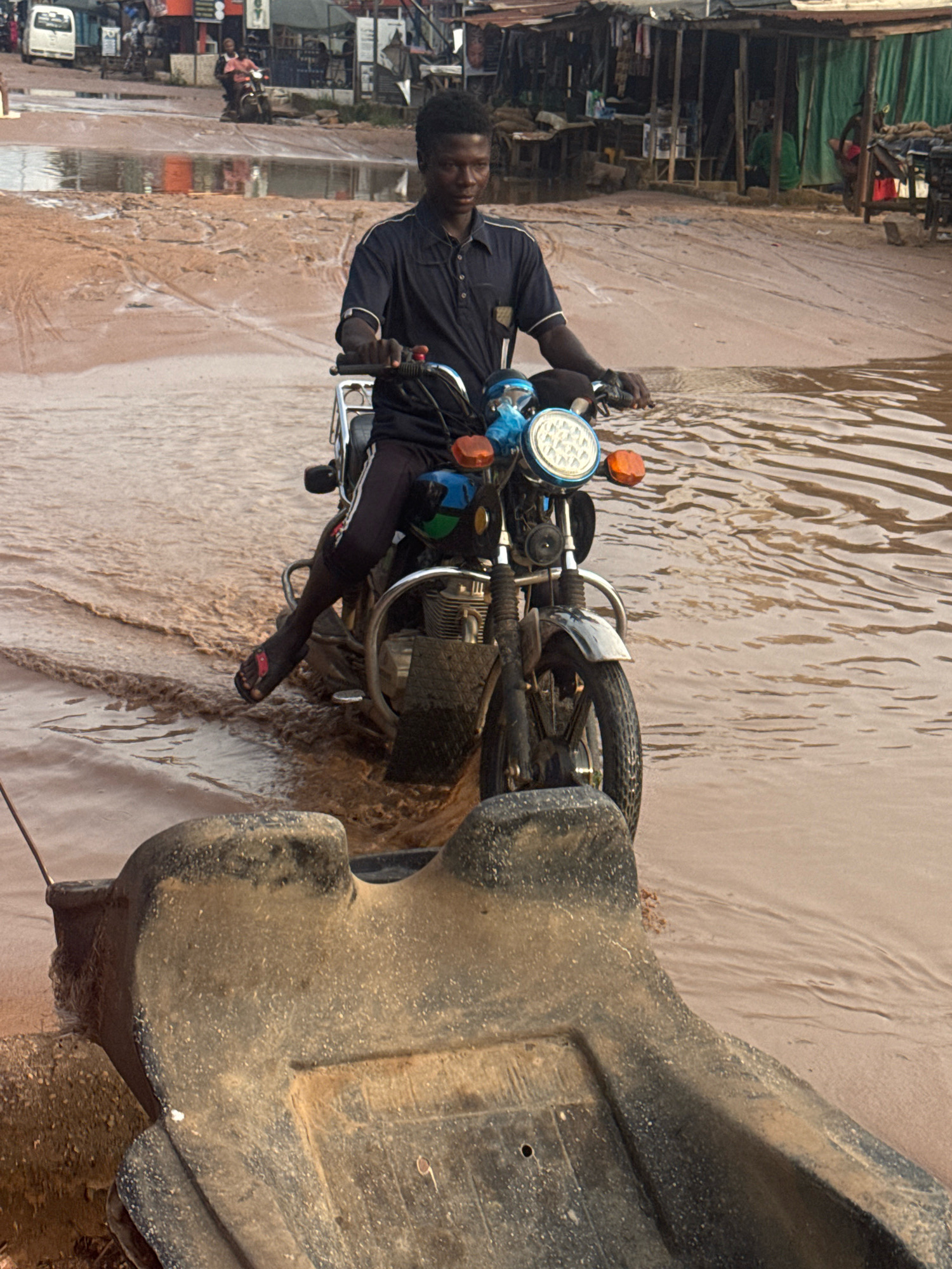 A person rides a motorcycle through flooded, muddy terrain in what appears to be the city of Ihiagwa, Nigeria. The person, dressed in a dark blue short-sleeved shirt and black pants with white stripes, is seated on the motorcycle and is looking slightly to the right. The motorcycle, a dark color, has a prominent round headlight with a blue frame and orange turn signals. The front wheel is submerged in the brown, murky water, and the motorcycle is creating ripples as it moves forward. In the foreground, a large, dark-colored object, possibly a boat or a large container, is partially submerged in the water, with its textured surface facing the camera. The background shows a street lined with makeshift stalls and structures, some with corrugated metal roofs. Several other motorcycles and a white van are visible further down the street. The ground is a mixture of mud and water, indicating recent rainfall or poor drainage.
