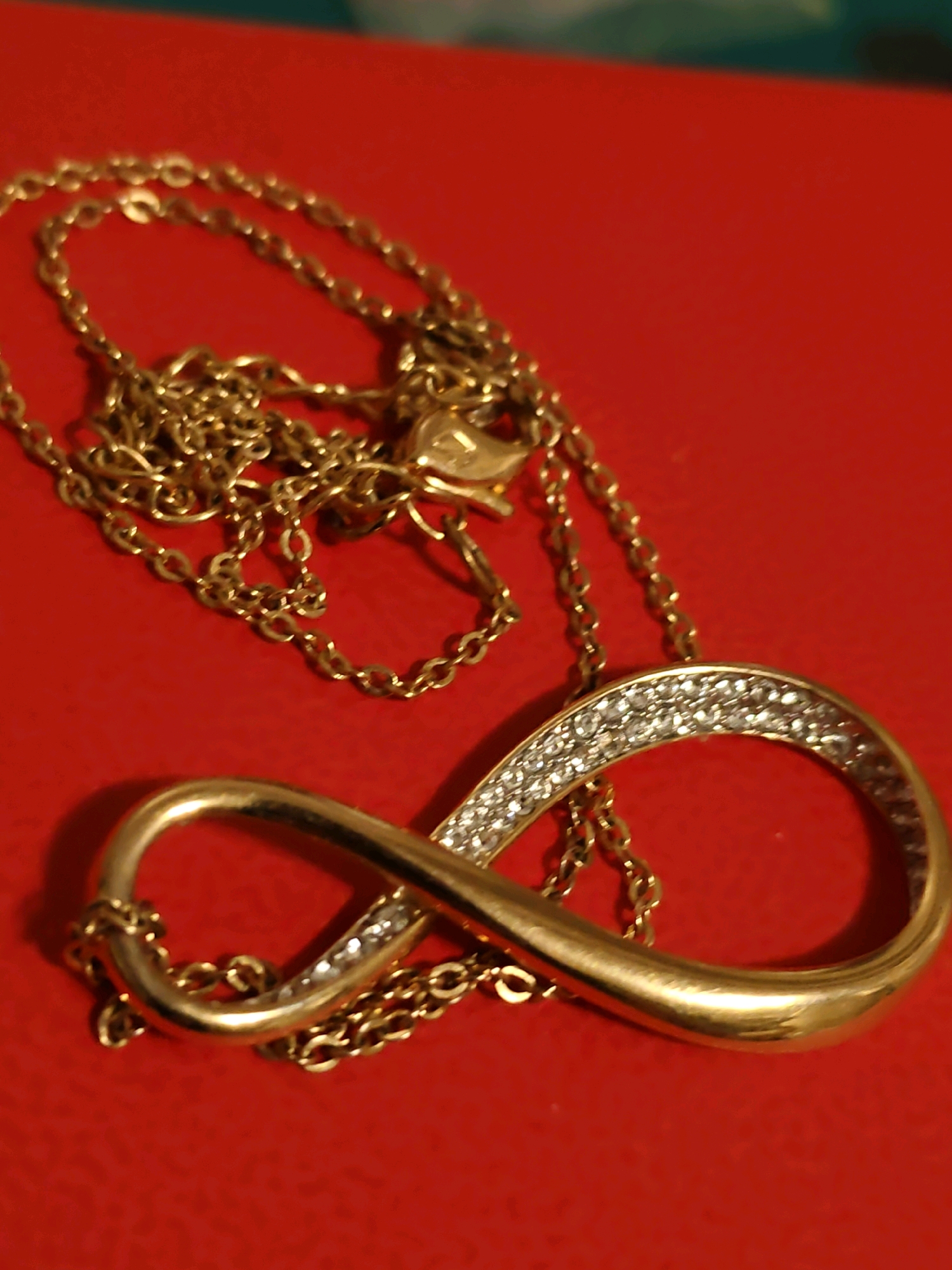 A close-up shot captures a gold infinity necklace with small, clear stones adorning one loop, resting on a vibrant red surface. The necklace is coiled and draped, with the pendant positioned prominently in the foreground. The background is a blurred red surface, suggesting a backdrop or table. The lighting is warm, casting subtle shadows. The image is sharply focused on the necklace. There is no information in the image that can be used to determine its location in the city of Rostov-on-Don, Russia.