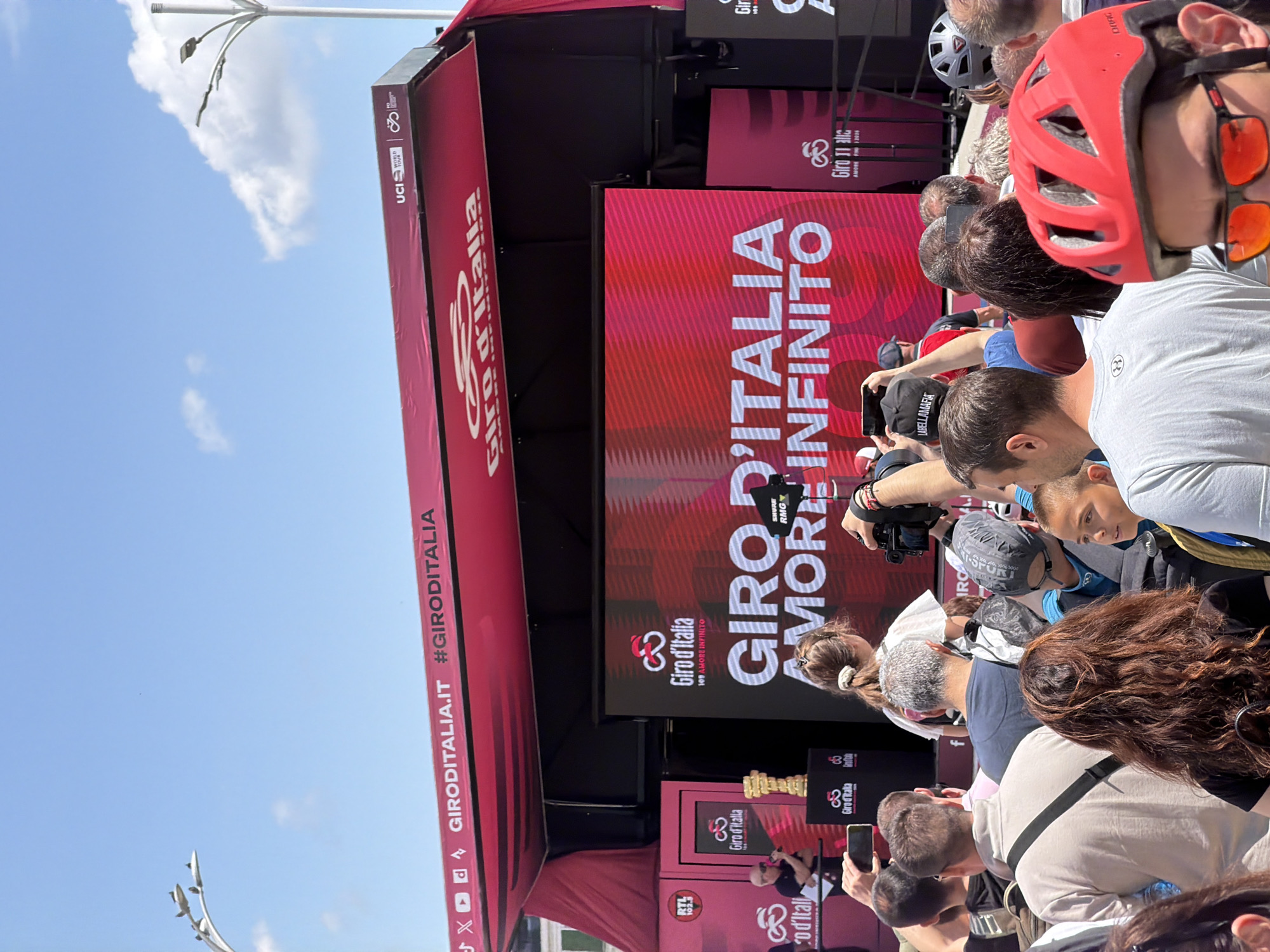 The image shows a crowd of people standing in front of a stage decorated for the Giro d'Italia. The stage has branding elements such as the Giro d'Italia logo and the phrase "GIRO D'ITALIA AMORE INFINITO." Several individuals in the crowd are holding phones, likely taking pictures or videos of an event occurring on stage. Some wear hats, and one person has a red cycling helmet with orange-tinted sunglasses. The background features a clear blue sky with some light clouds, and a light pole is visible. The location is not specified in the image, therefore I cannot assume the event to be taking place in Plovdiv, Bulgaria.