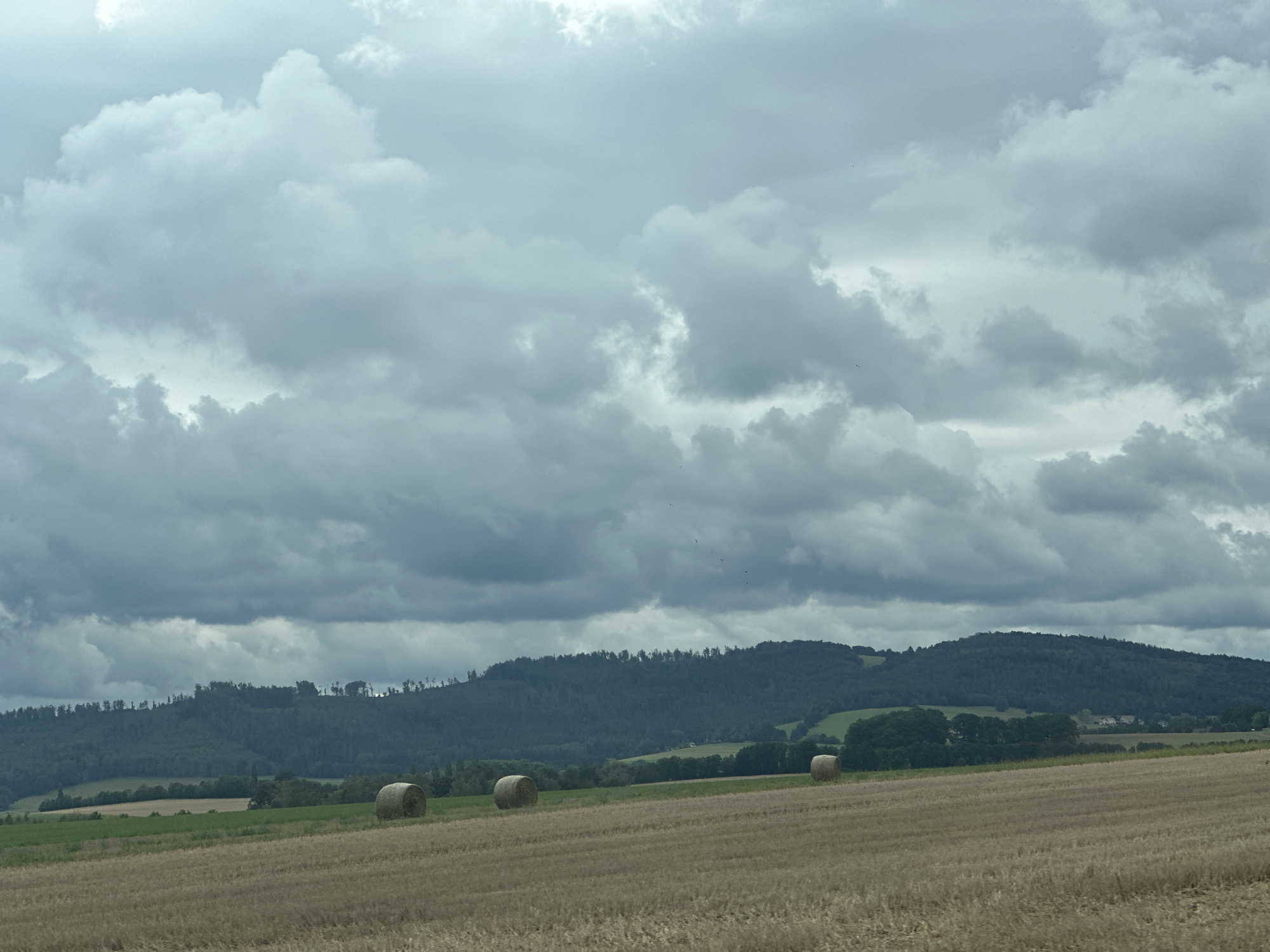 The image depicts a rural landscape under a cloudy sky. In the foreground, a mowed field, likely containing stubble from a recent harvest, stretches horizontally across the frame. Three large, round hay bales are scattered across this field. The middle bale is positioned slightly further back and to the left of the other two. Beyond the field, a gently sloping hill is covered in dark green trees, forming a dense forest that extends to the horizon. In the distance, atop the tree line, a few buildings are visible, suggesting a settlement nestled within the landscape. The sky is filled with heavy, grey clouds, indicating overcast weather. This scene is located in Großpostwitz, Germany.