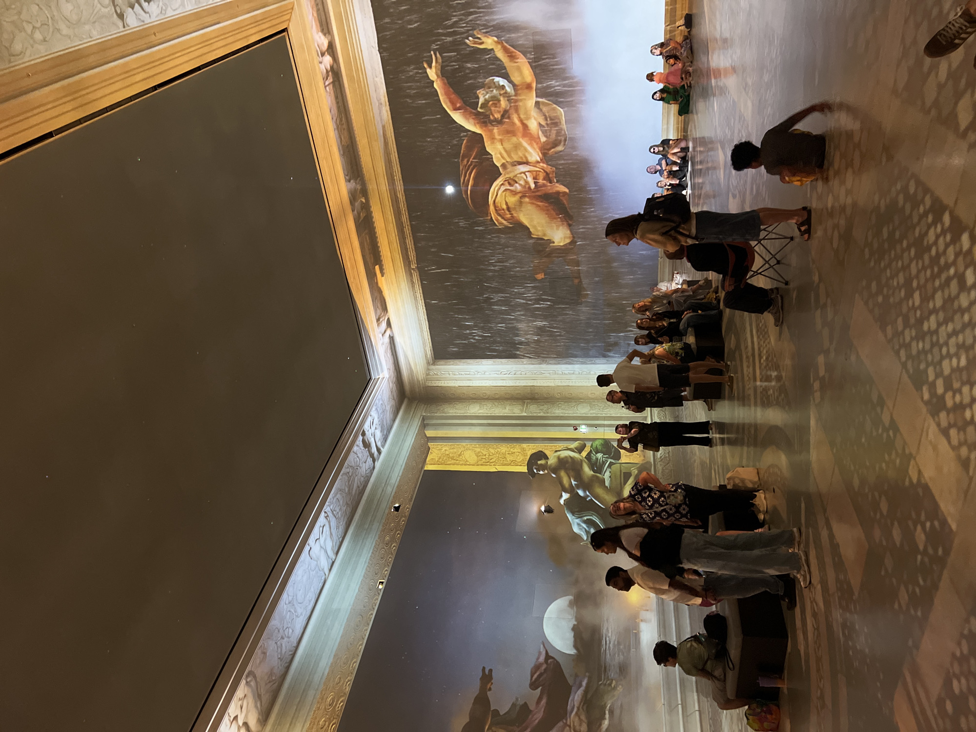 The image depicts an immersive art exhibition within a large, ornate hall. The walls are adorned with projected artwork, one side featuring a celestial scene with a large moon and figures, and the other side displaying a dynamic projection of a falling or flying figure amidst what appears to be rain. A diverse group of people are observing the projections, some seated on benches and chairs, others standing. The floor is polished and reflects the projected images and overhead lighting, creating intricate shadow patterns. The architecture suggests a historic building, possibly a museum or gallery. The setting is dimly lit to enhance the visual impact of the projections. The individuals are dressed in casual attire, indicating a public exhibition. The overall impression is one of awe and engagement with the large-scale digital art installation.
