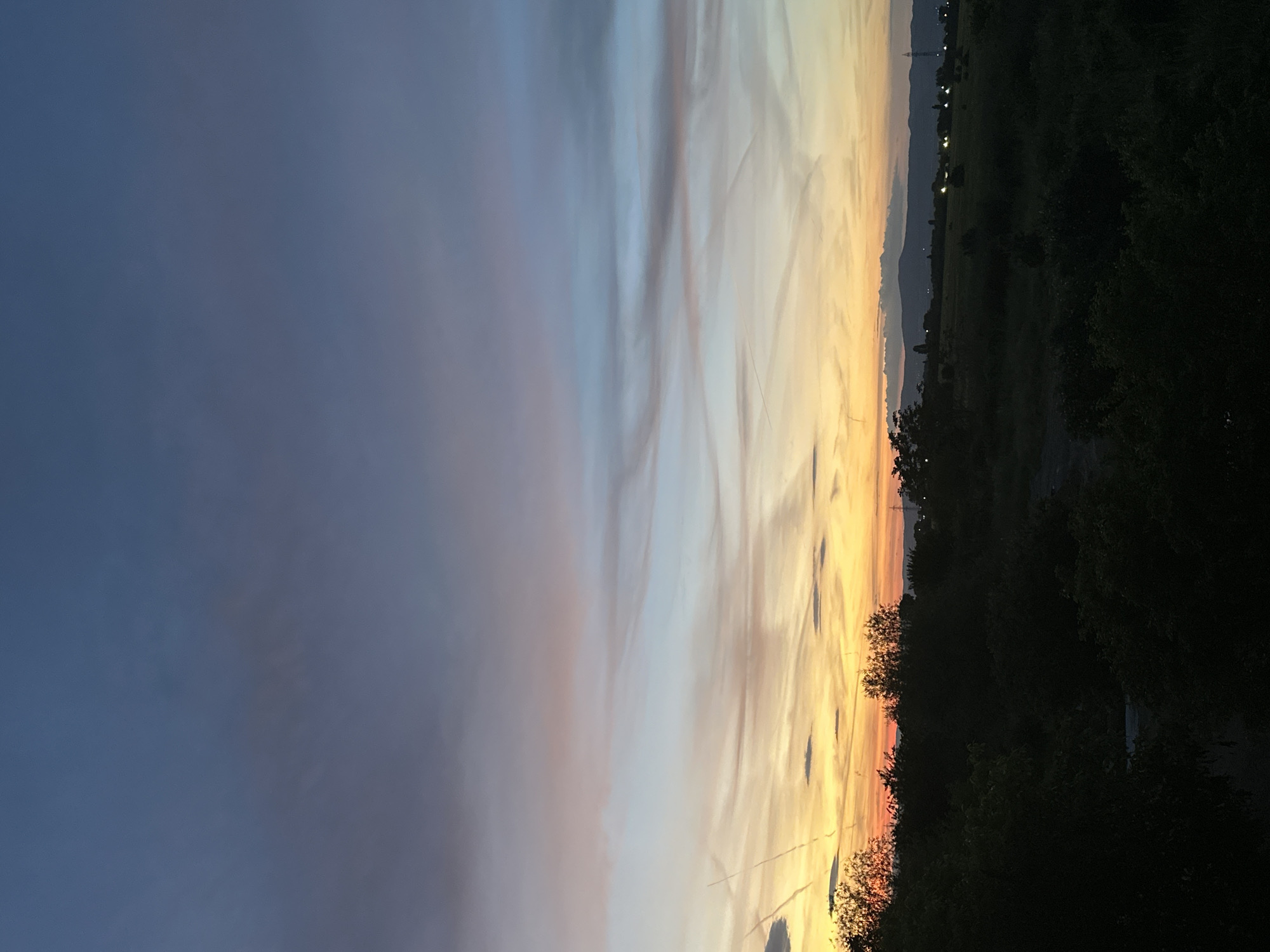 The image shows a twilight or sunset landscape. The sky takes up a large portion of the frame, transitioning from dark blue on the left to orange and yellow hues on the right, with visible contrails crisscrossing the sky. The horizon line, slightly obscured, features dark foliage and some indistinct structures or buildings. Due to the low light and limited detail, it's not possible to definitively identify the specific location in Sofia, Bulgaria.