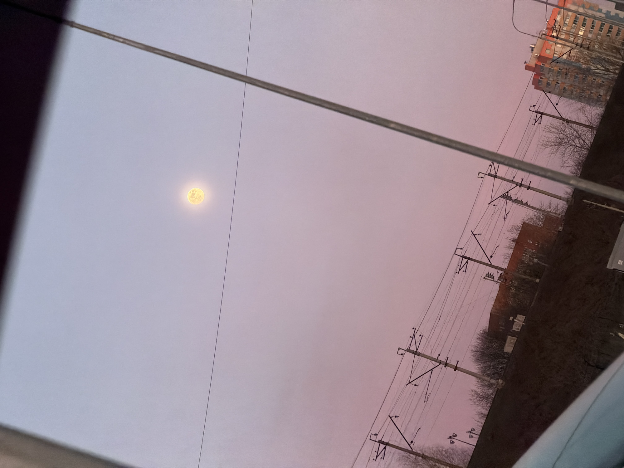 The image shows an urban scene in Perm, Russia, taken at dusk or dawn. The sky is a mix of light purple and pink hues. A full moon is visible in the left side of the frame. The landscape is dominated by utility poles and power lines that traverse the sky. In the background, there are buildings, including a tall apartment complex with orange accents. Some bare trees are interspersed among the buildings. The photo appears to have been taken from inside a vehicle, with a portion of the car's interior visible in the bottom corner and parts of the window frame visible.