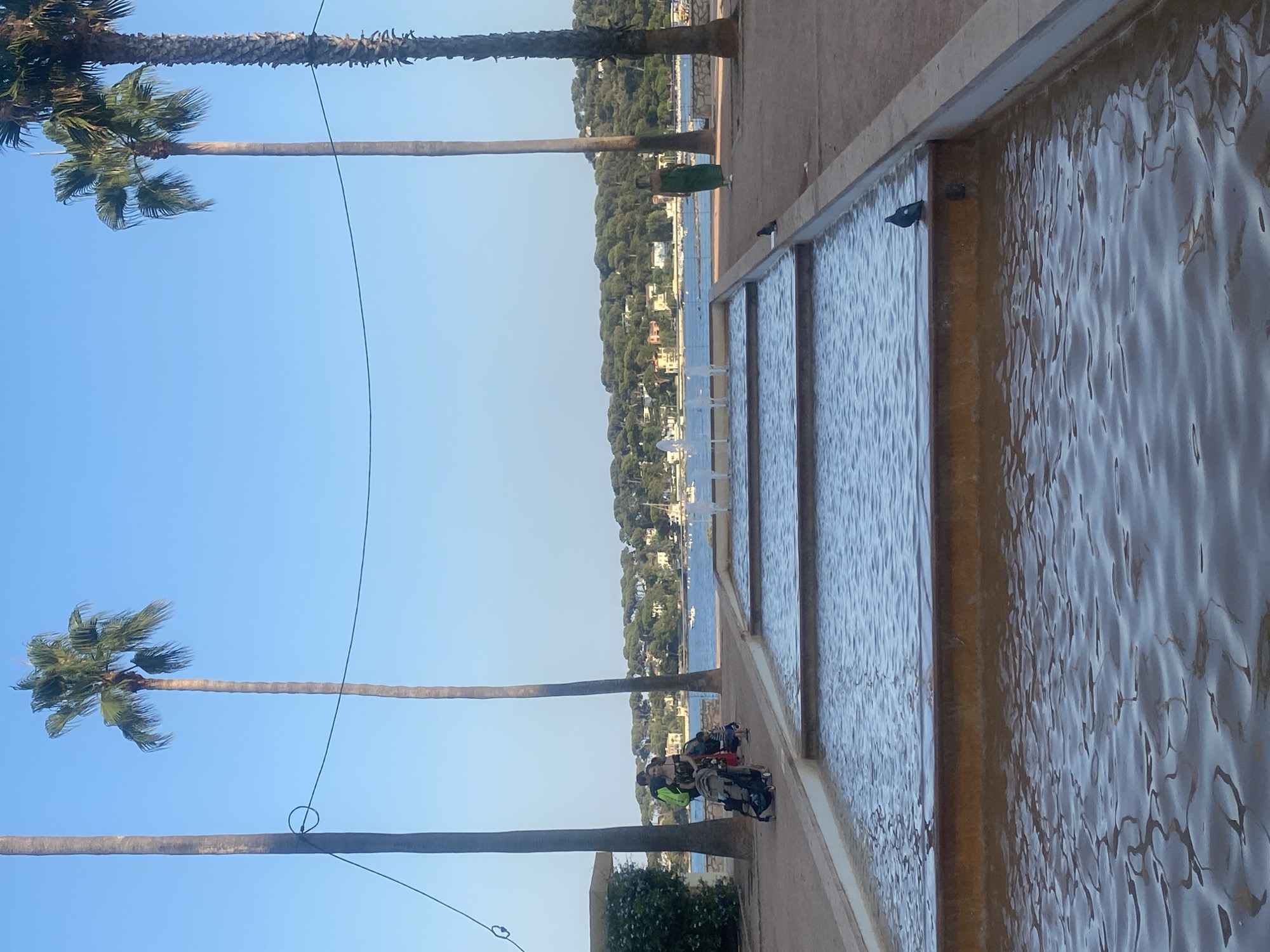 A sunny afternoon scene is captured along the waterfront in Antibes, France. In the foreground, a tiered water feature with shallow pools reflects the blue sky. Palm trees, characteristic of the Mediterranean climate, are prominent throughout the image, with two tall specimens dominating the right side of the frame and another on the left.
A family is visible in the middle ground, with a man and woman pushing a stroller. A young child stands near them. To the right, a woman in a green dress walks along the paved walkway. A pigeon is perched on the edge of one of the water pools, and another is visible on the walkway.
Beyond the water feature, the Antibes coastline unfolds with a bay dotted with sailboats. The backdrop consists of a verdant hillside covered in trees and dotted with houses, suggesting a residential area overlooking the sea. The sky is a clear, vibrant blue, and the overall impression is one of a peaceful, picturesque coastal town.