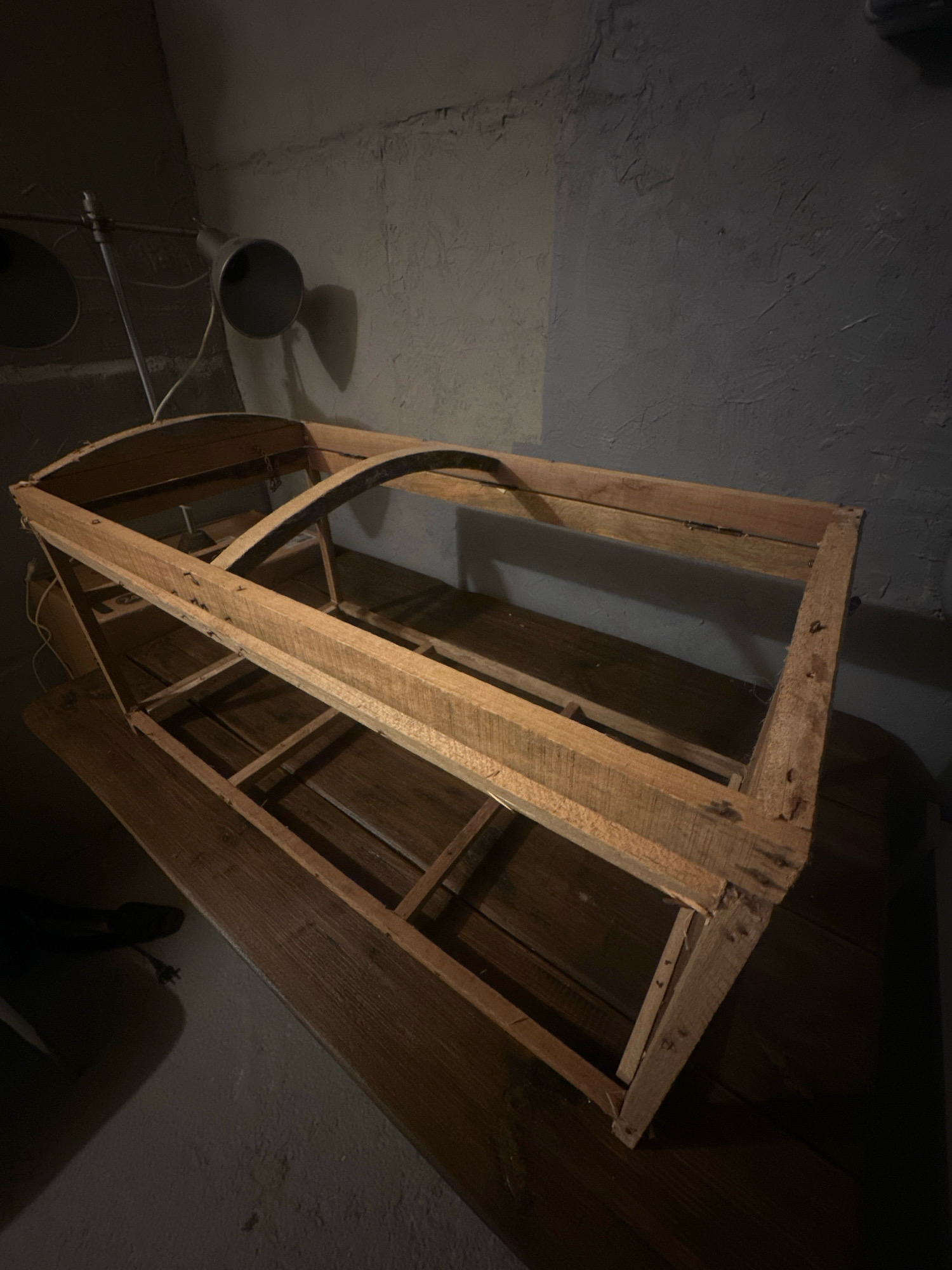 The image shows a wooden shelf structure on a dark wooden surface, likely a table. The shelf appears to be unfinished and made of rough-hewn planks. It has two tiers, with the top tier featuring a curved wooden element. The structure is illuminated by a directional light source, casting shadows on the textured grey wall behind it. A metallic pipe with a light fixture is mounted on the wall above and to the left of the shelf. The overall scene suggests an indoor, possibly workshop or storage area, environment. The location is identified as Lapės, Lithuania. There are no people visible in the image.