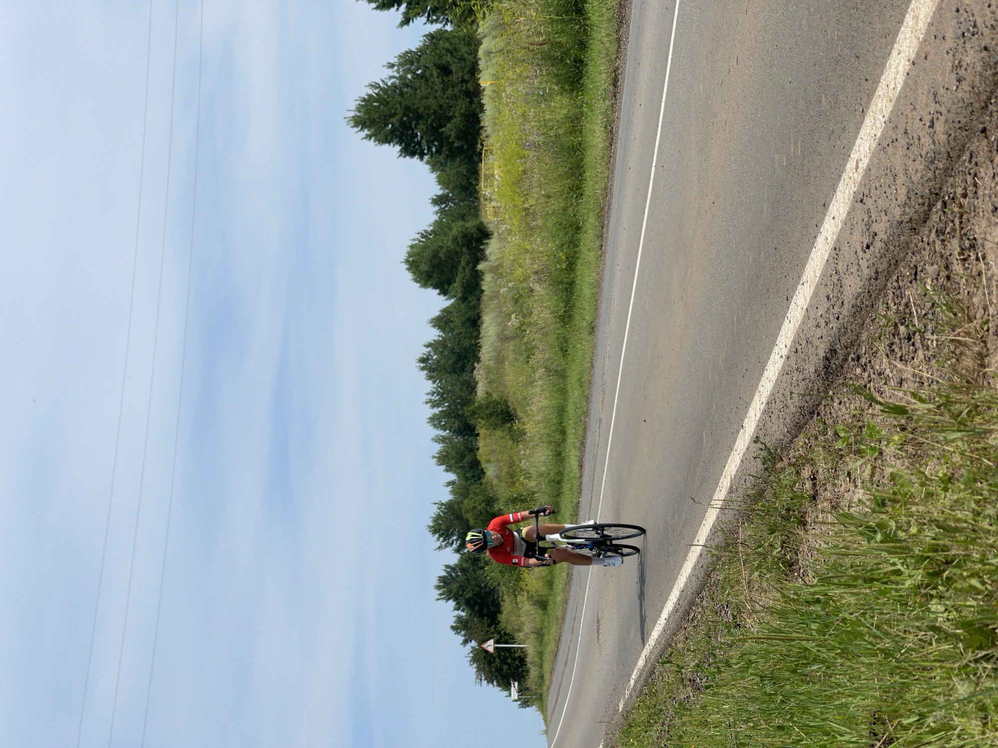 A cyclist wearing a red and white jersey, black shorts, a yellow and green helmet, and sunglasses rides a white road bicycle on an asphalt road. The road has a white line marking its edge and another line down its center. The cyclist is on the right side of the road, heading towards the viewer. The background consists of a grassy verge with wildflowers and trees, and a clear blue sky with some thin clouds and power lines. A triangular road sign with a symbol is visible on the left side of the road, with text on it that is partially obscured but appears to be in Russian. The overall scene depicts an outdoor cycling activity on a rural road. The location is identified as Perevoznoe, Russia.