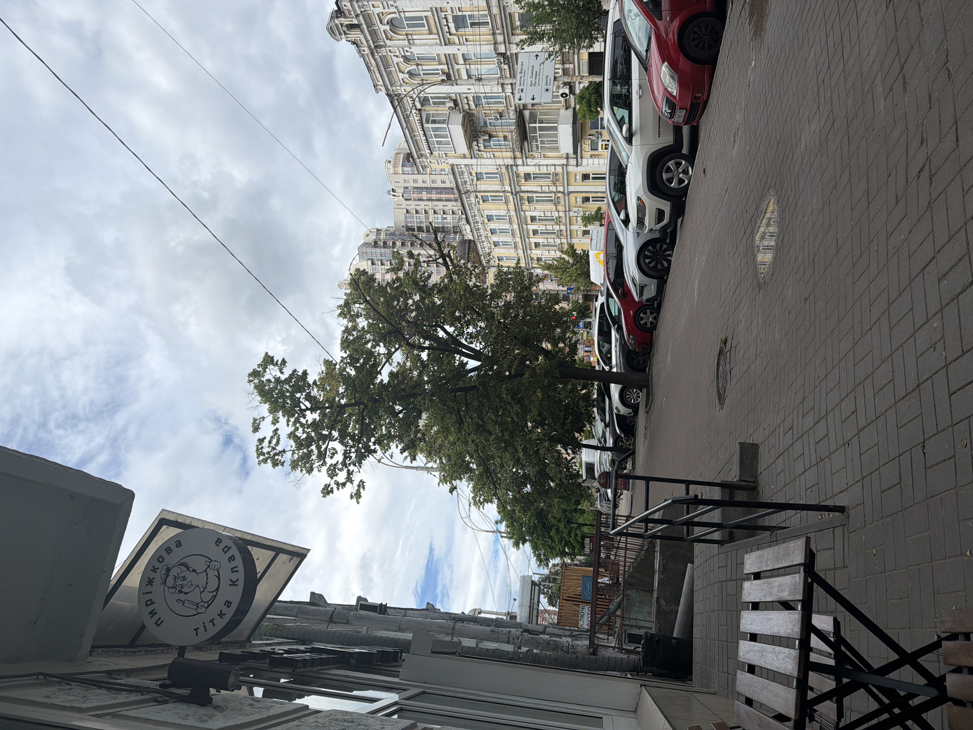 The image depicts a street scene in Kyiv, Ukraine, with a focus on a row of parked cars, a building with traditional architecture, and a tree. The cars, including a red, white, and black one, are parked along a brick-paved sidewalk. In the background, a multi-story building showcases ornate architectural details, with balconies and numerous windows. A large tree with green foliage stands prominently, its branches extending towards the sky.
Above the scene, the sky is filled with a mix of clouds and patches of blue, suggesting it might be a partly cloudy day. The lighting indicates it's daytime. There is a sign visible on the left side of the image, round in shape with a logo of a pig and text. The text on the sign appears to be in Cyrillic script. Further down the street, another sign with directional arrows and Cyrillic text is attached to the building.
The overall impression is a typical urban street in a European city, with a blend of older and perhaps more modern architectural styles. There are no people clearly visible in the image, and the scene appears static, focusing on the environment rather than an active event. The presence of parked cars and sidewalk seating suggests this could be a commercial or residential street.