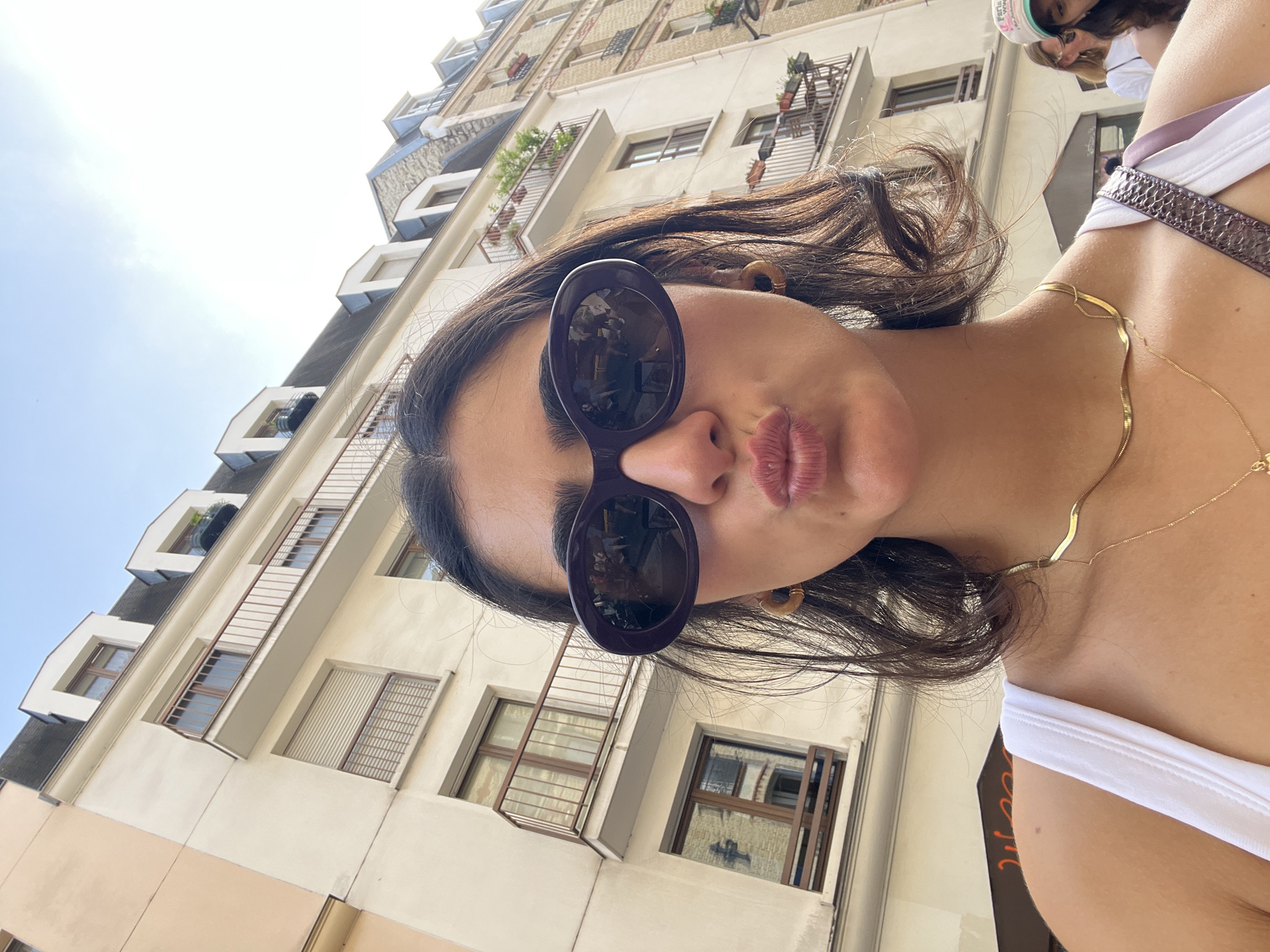 A low-angle shot depicts a woman in a white tank top, gold necklace, and oversized sunglasses. She has dark hair and is puckering her lips towards the camera. Behind her is a light-colored building with multiple windows and small balconies decorated with plants. The shot appears to be taken in Paris, France, based on the architectural style of the building.